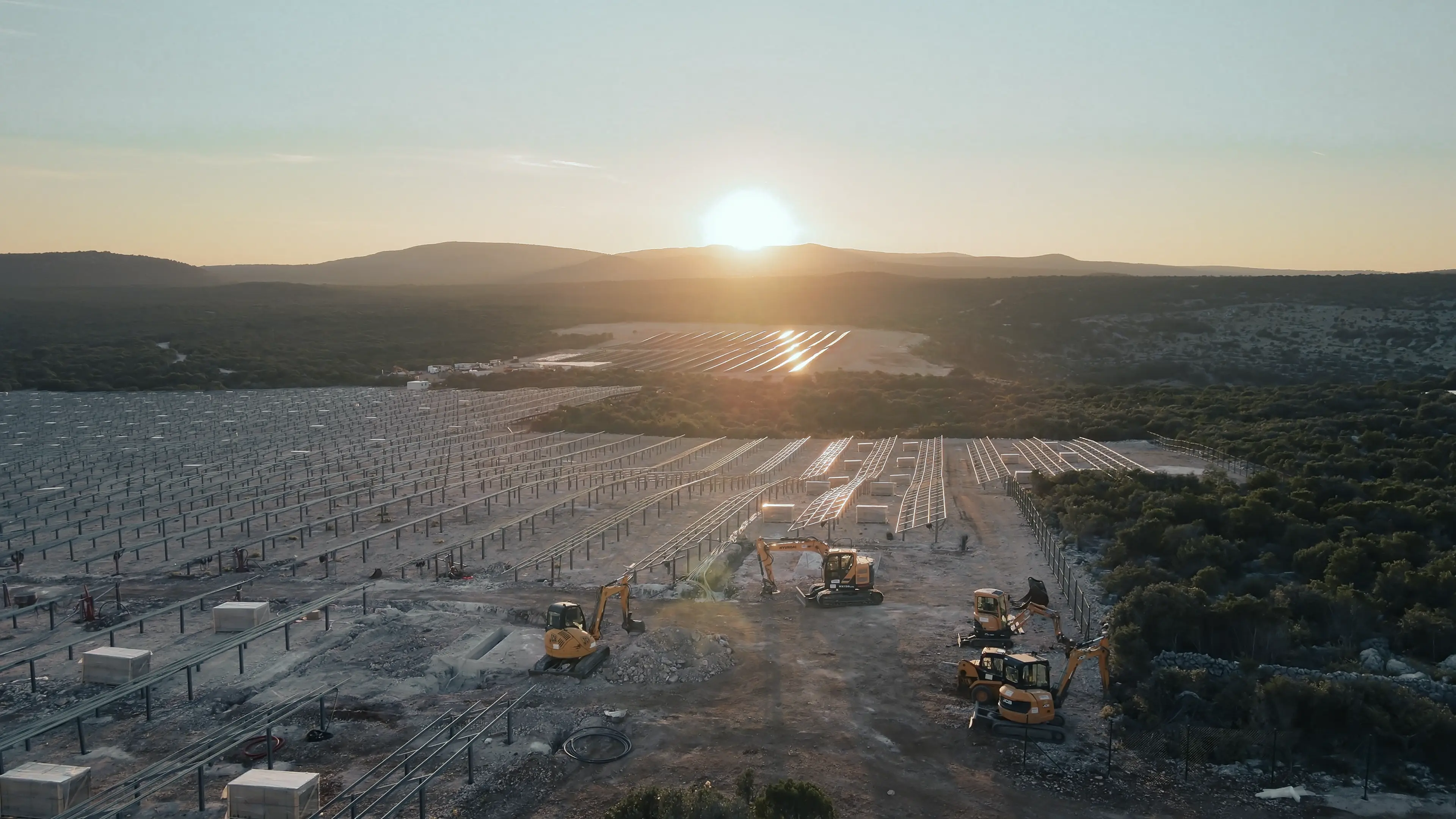 Sun setting over a large, rural solar farm, with construction vehicles on site and mountains in the distance.
