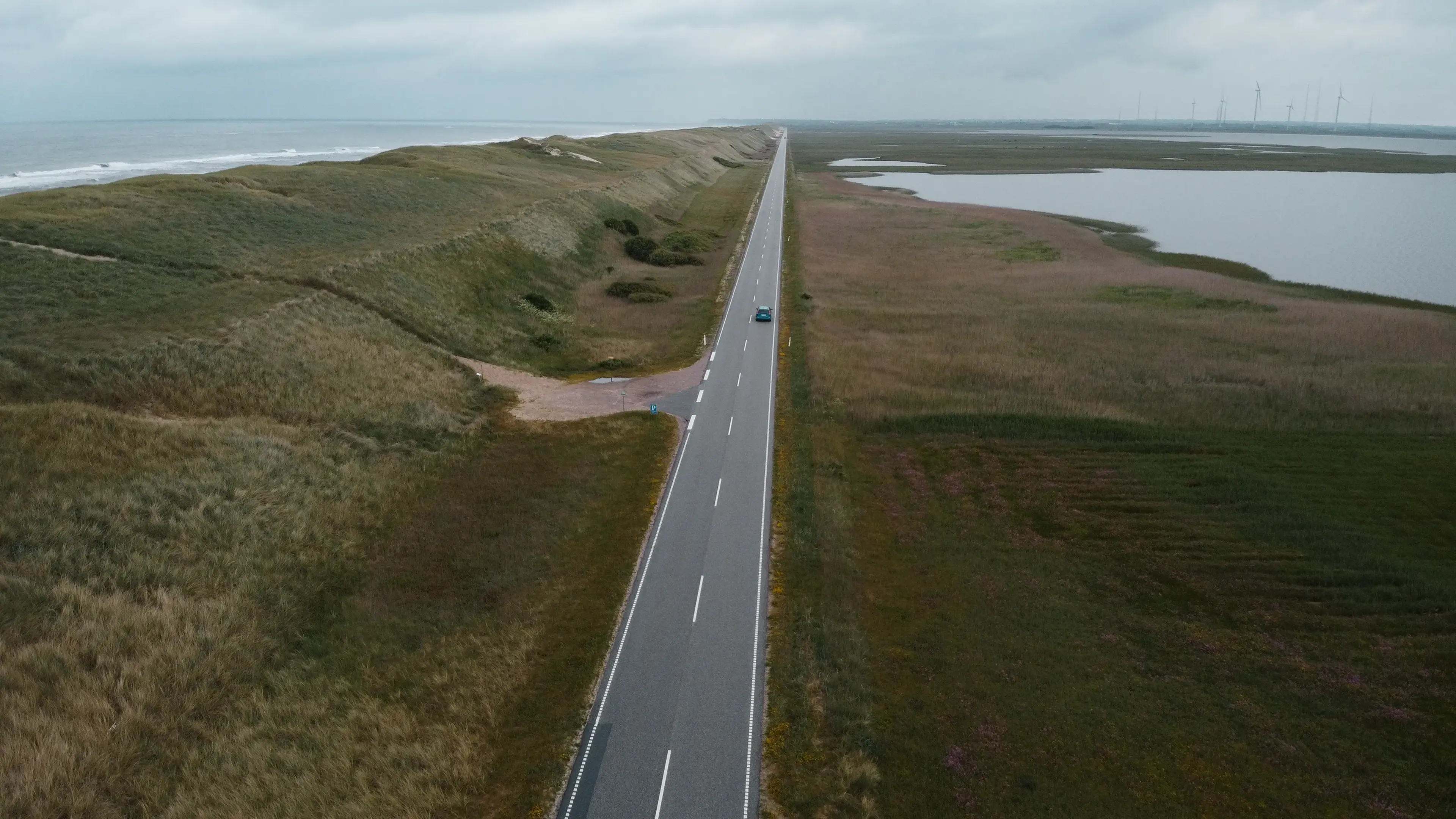 Straight road between grassy dunes and a lake on the right, a car driving along; wind turbines visible in the distance.