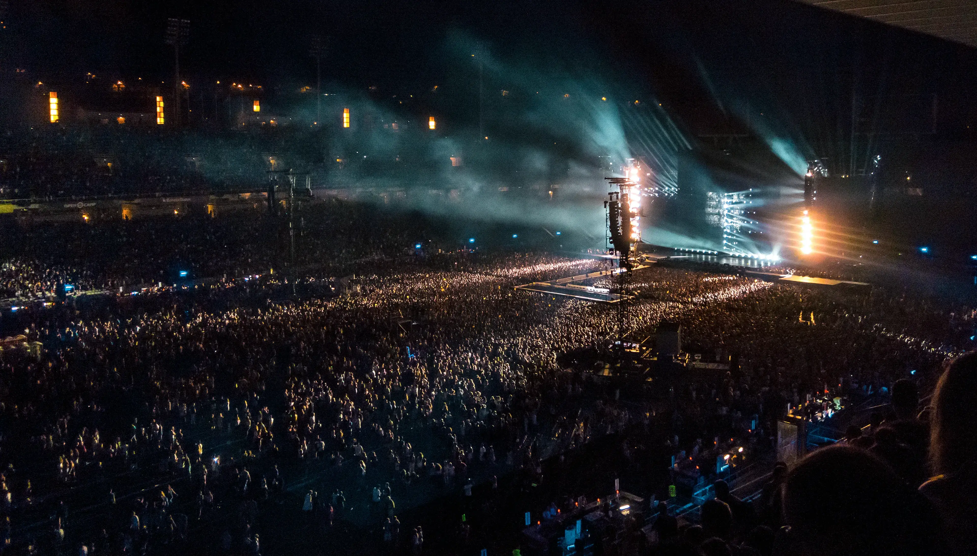 A large crowd attends a nighttime concert in a stadium, with bright stage lights and beams of light cutting through the smoke-filled air.