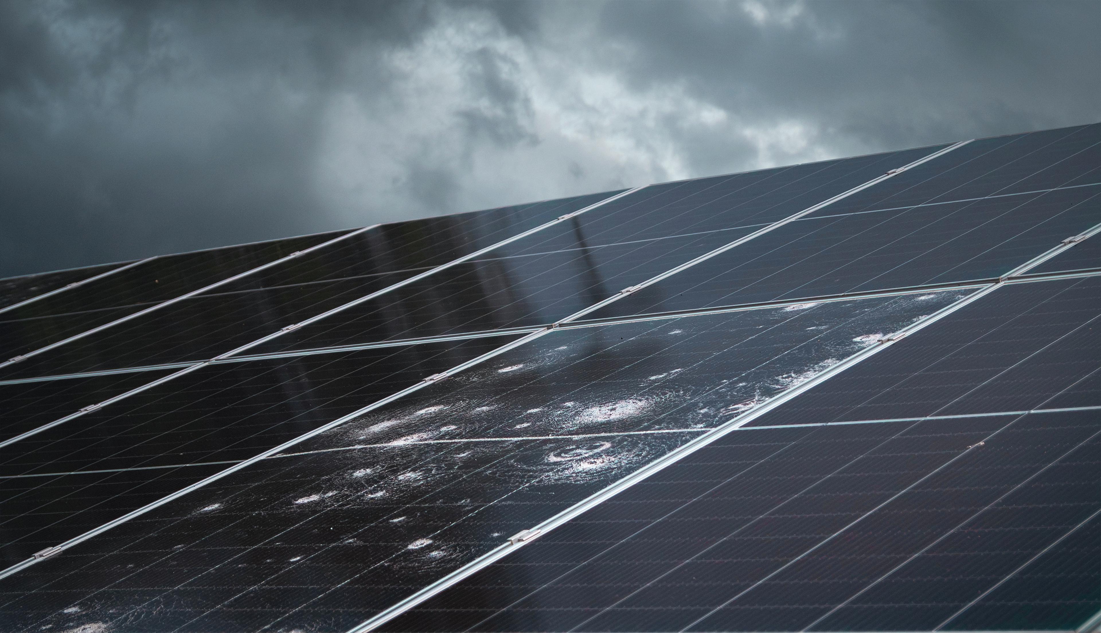 Close-up of solar panels broken by hail against a cloudy sky, reflecting dim light.