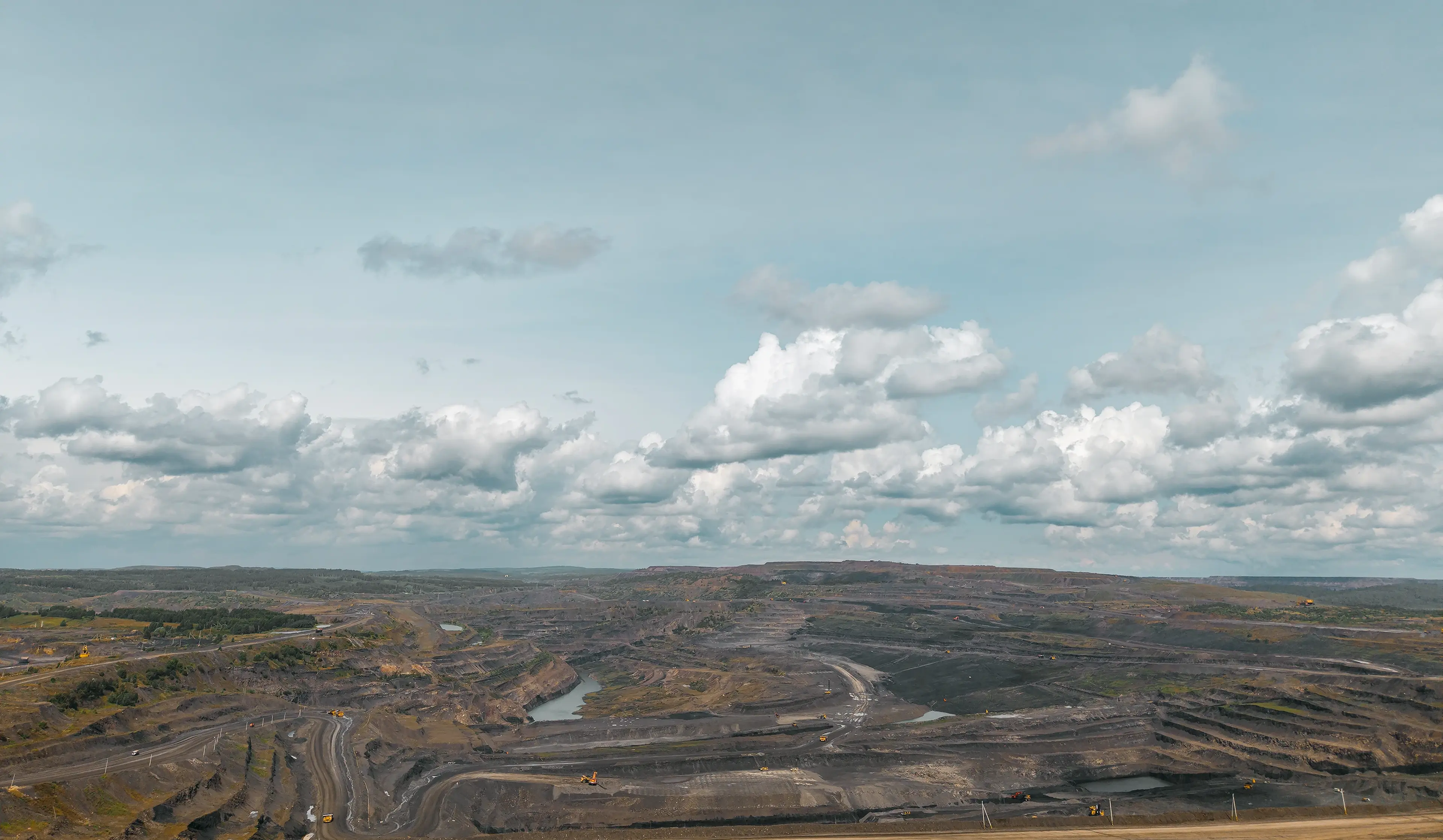 Expansive open-pit mine under a vast, cloudy sky, with winding roads and scattered mining equipment visible across the barren landscape.