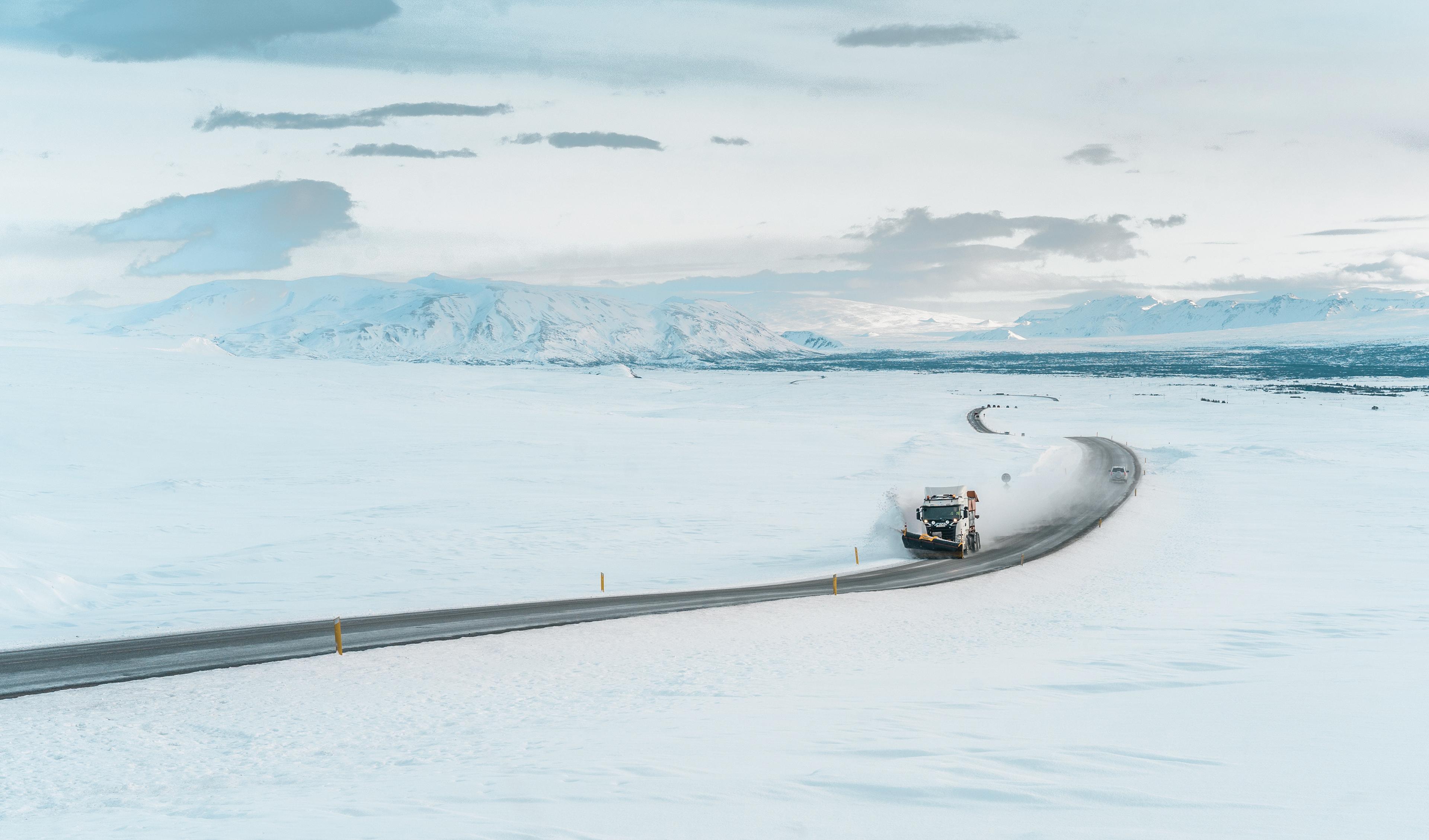A snowplow clears a winding road amidst a vast snowy landscape with distant mountains under a cloudy sky.