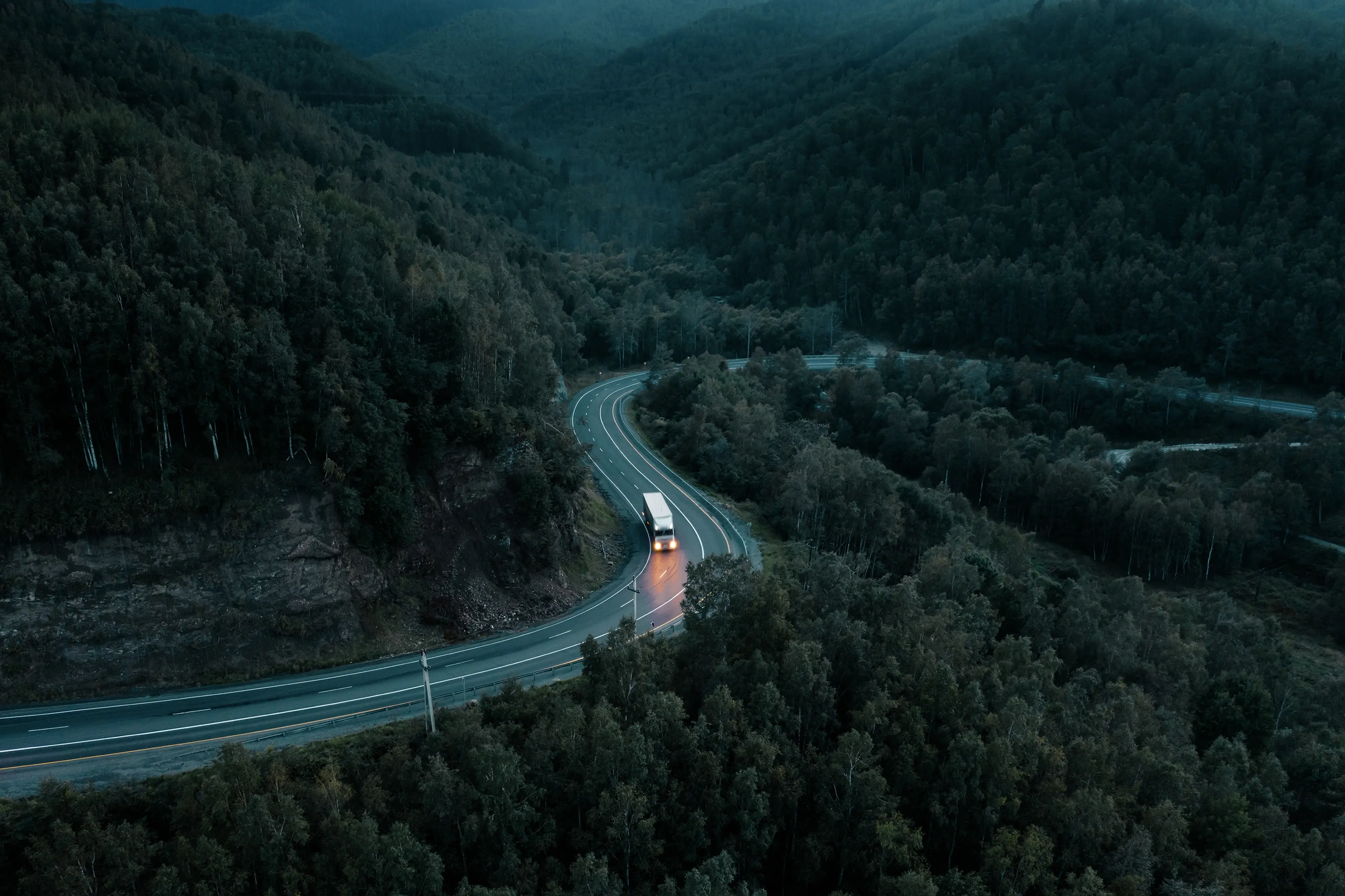 Aerial view of a truck on a winding road through a dense, mountainous forest at dusk, with surrounding lush greenery and dimly lit atmosphere.