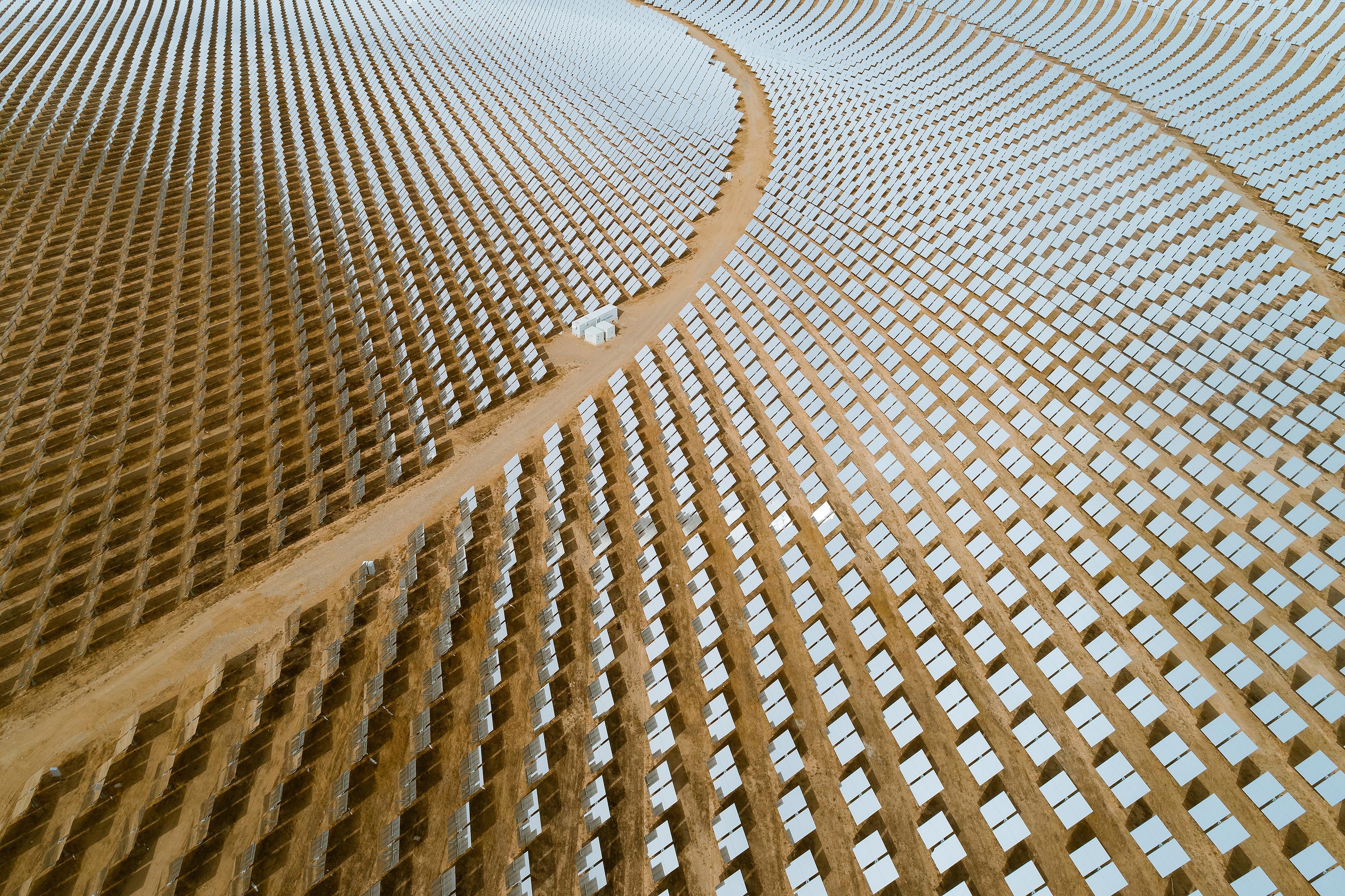 Aerial view of vast solar panel field with rows of panels on sandy terrain, intersected by a curved dirt path.