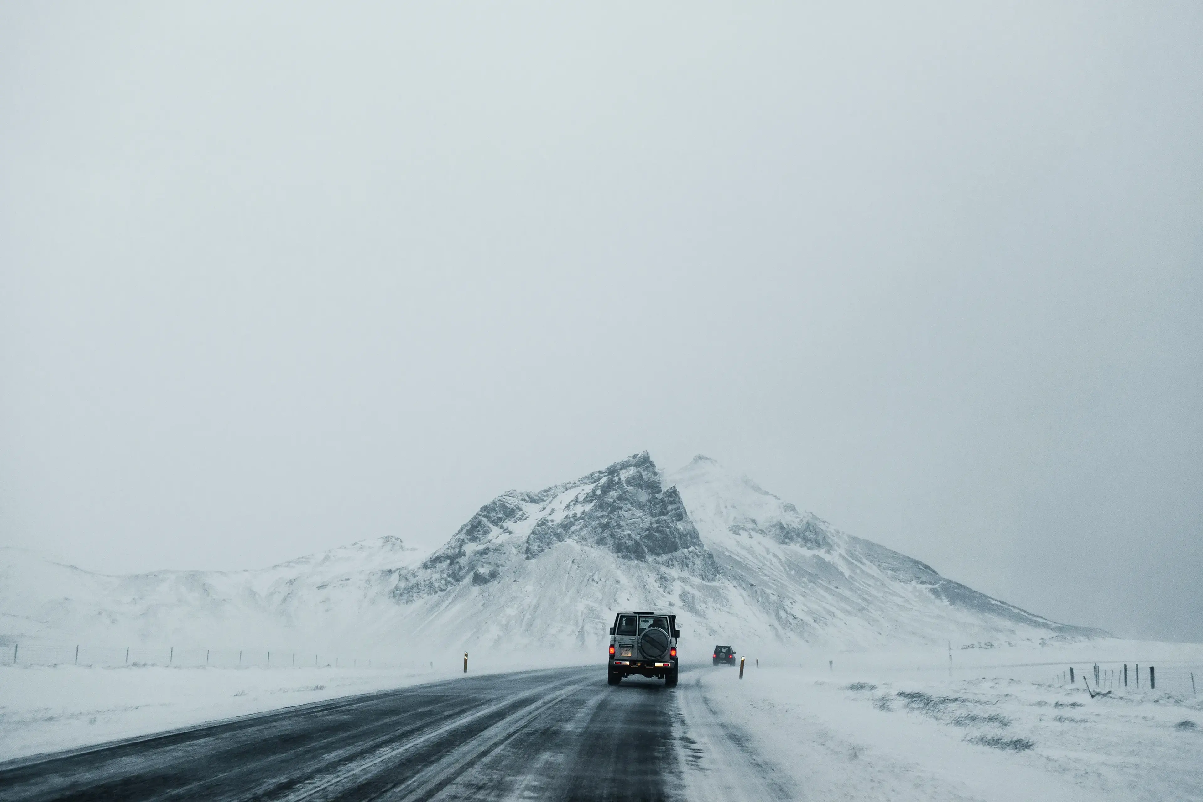 A car drives along a snowy road towards a distant, snow-covered mountain under an overcast sky.