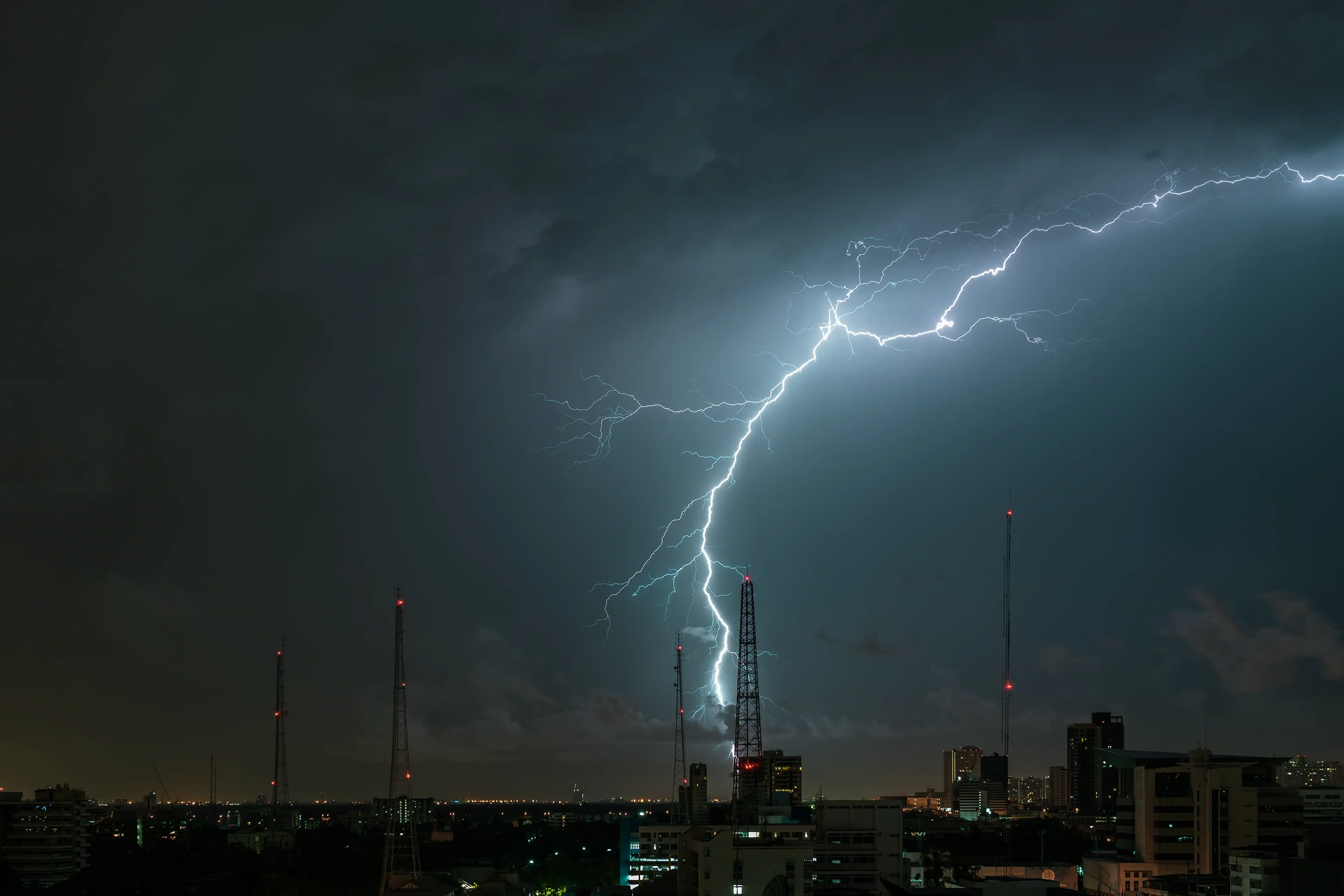 A dramatic bolt of lightning illuminates a dark, stormy night sky over a cityscape with prominent communication towers.