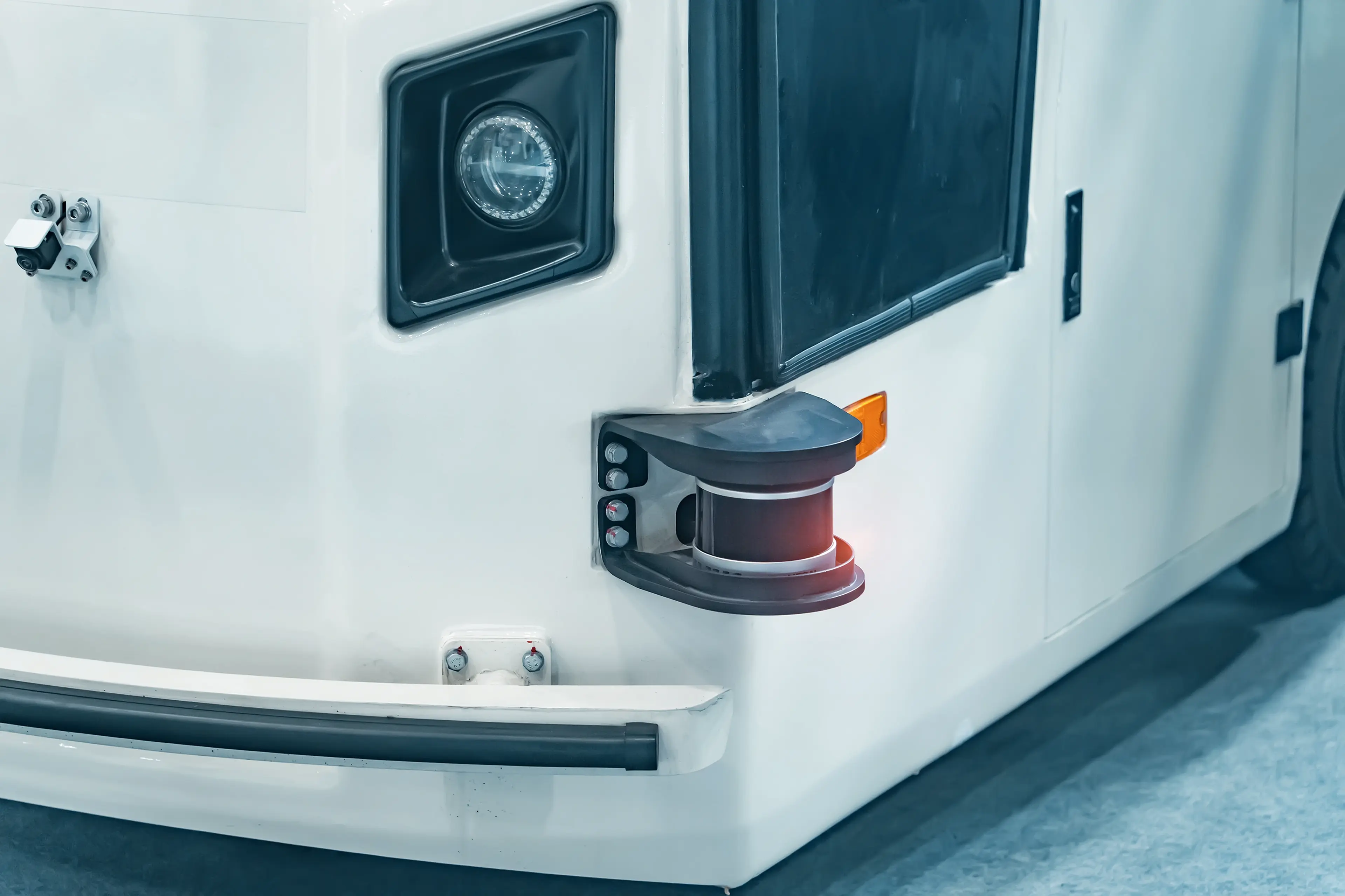 Close-up of a white vehicle with a front-mounted sensor or lidar system, featuring a headlight, black trim, and an amber side reflector.