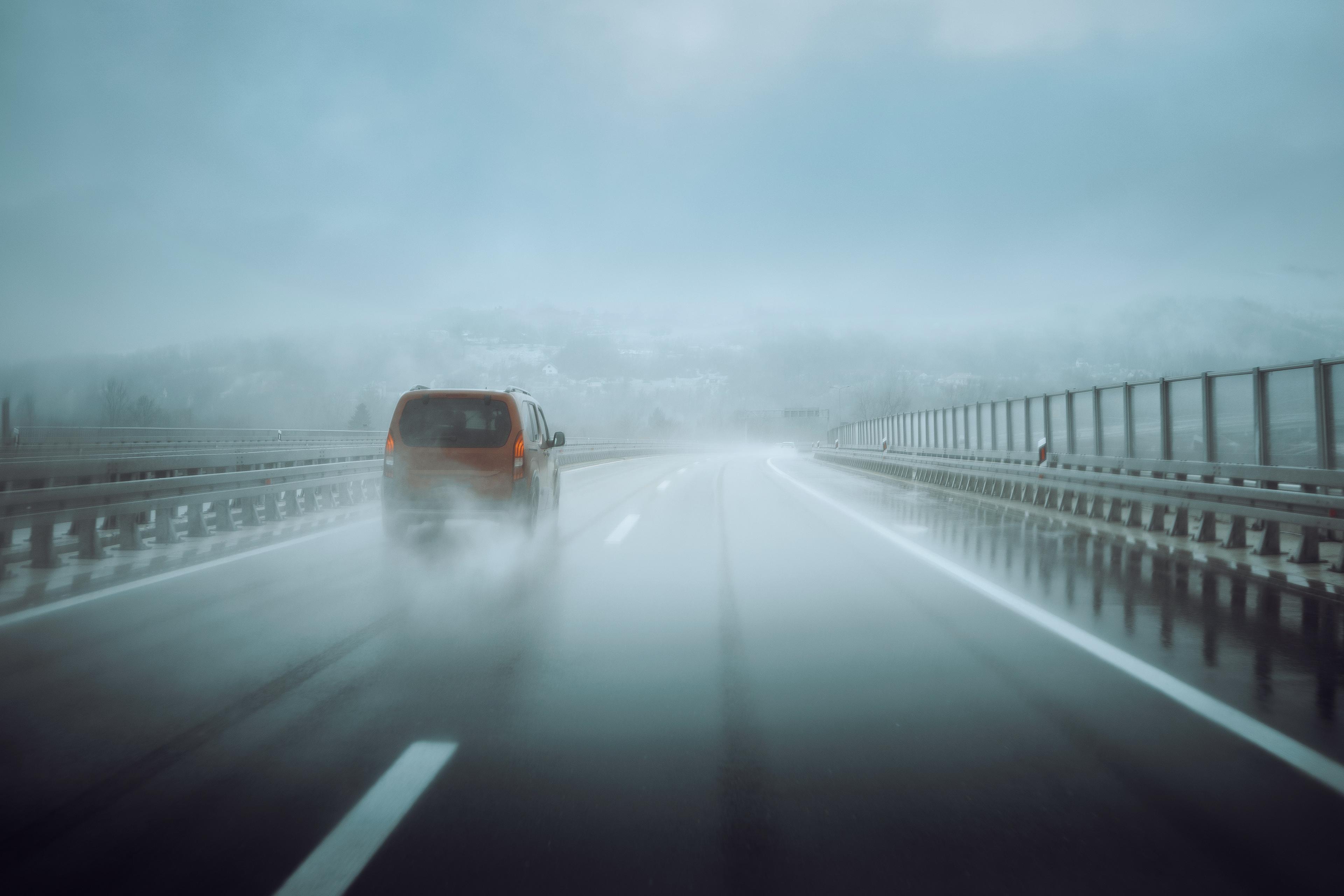 A red car drives on a wet, foggy highway, leaving a trail of mist. The scene is overcast, with blurred mountains faintly visible in the background.