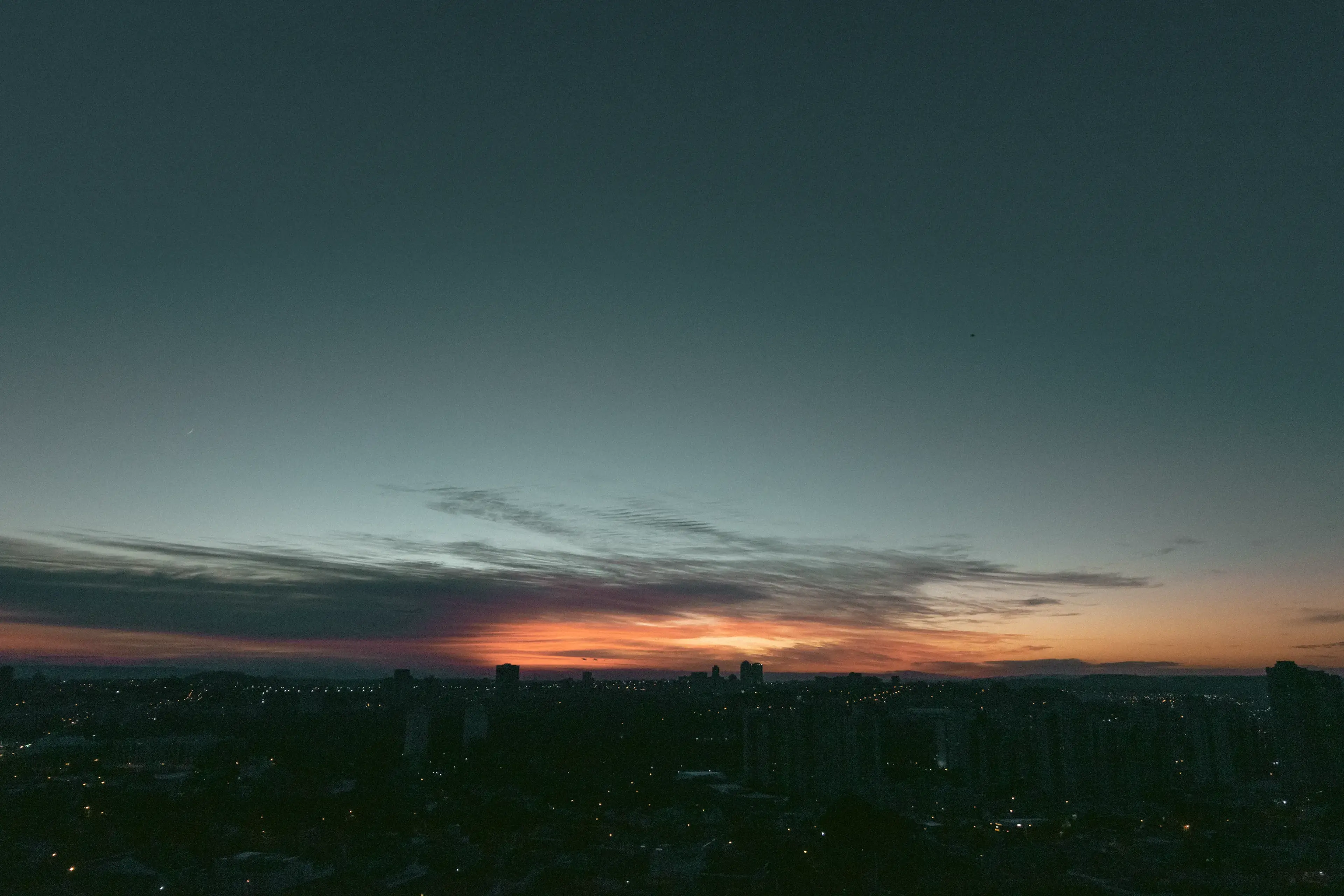 Cityscape at dusk with a vibrant gradient sunset; dark silhouetted buildings and soft, colorful clouds create a serene atmosphere.