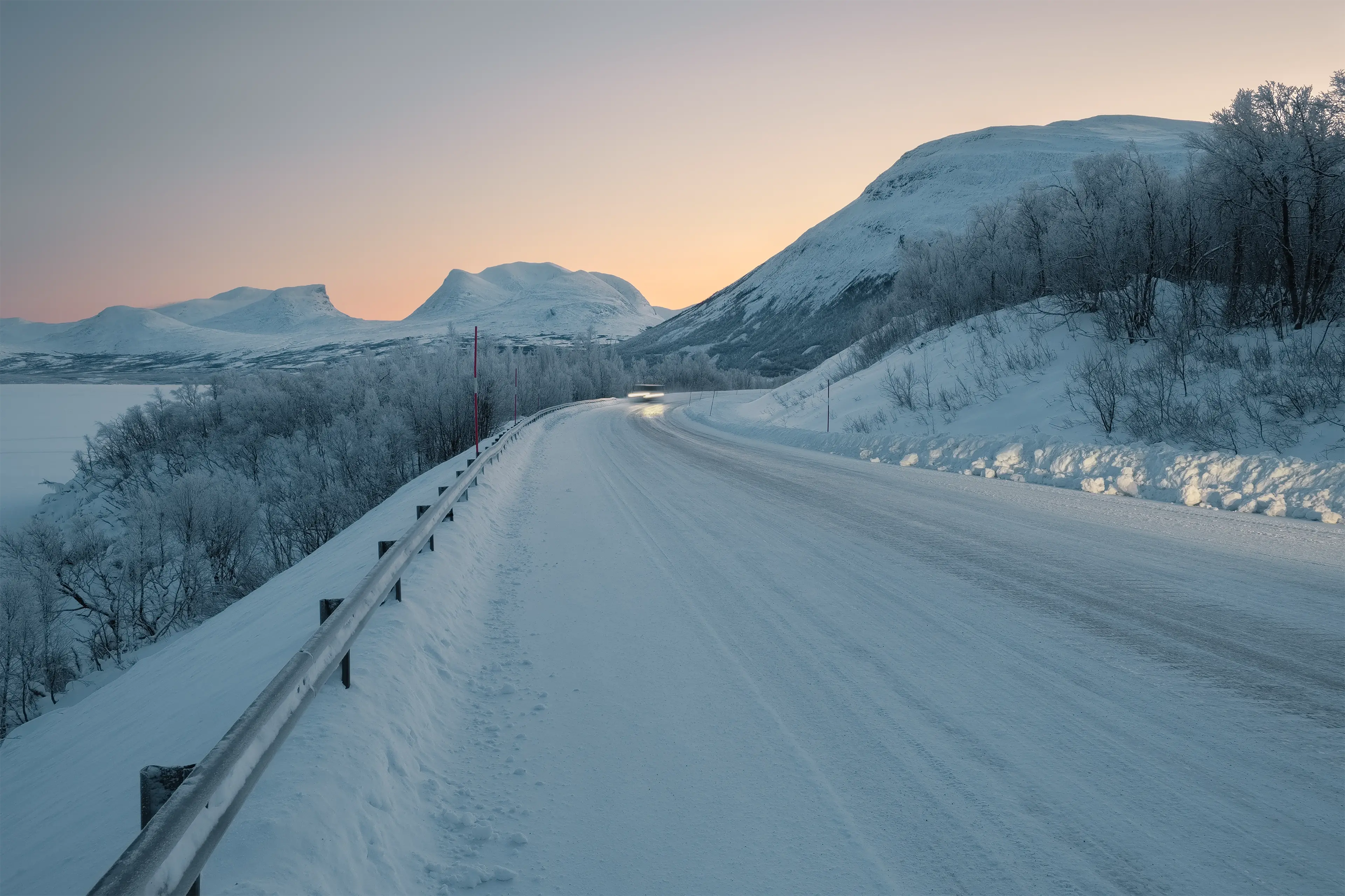 Snow-covered road winding through a winter landscape with snowy mountains and a soft pink sunrise sky in the background.