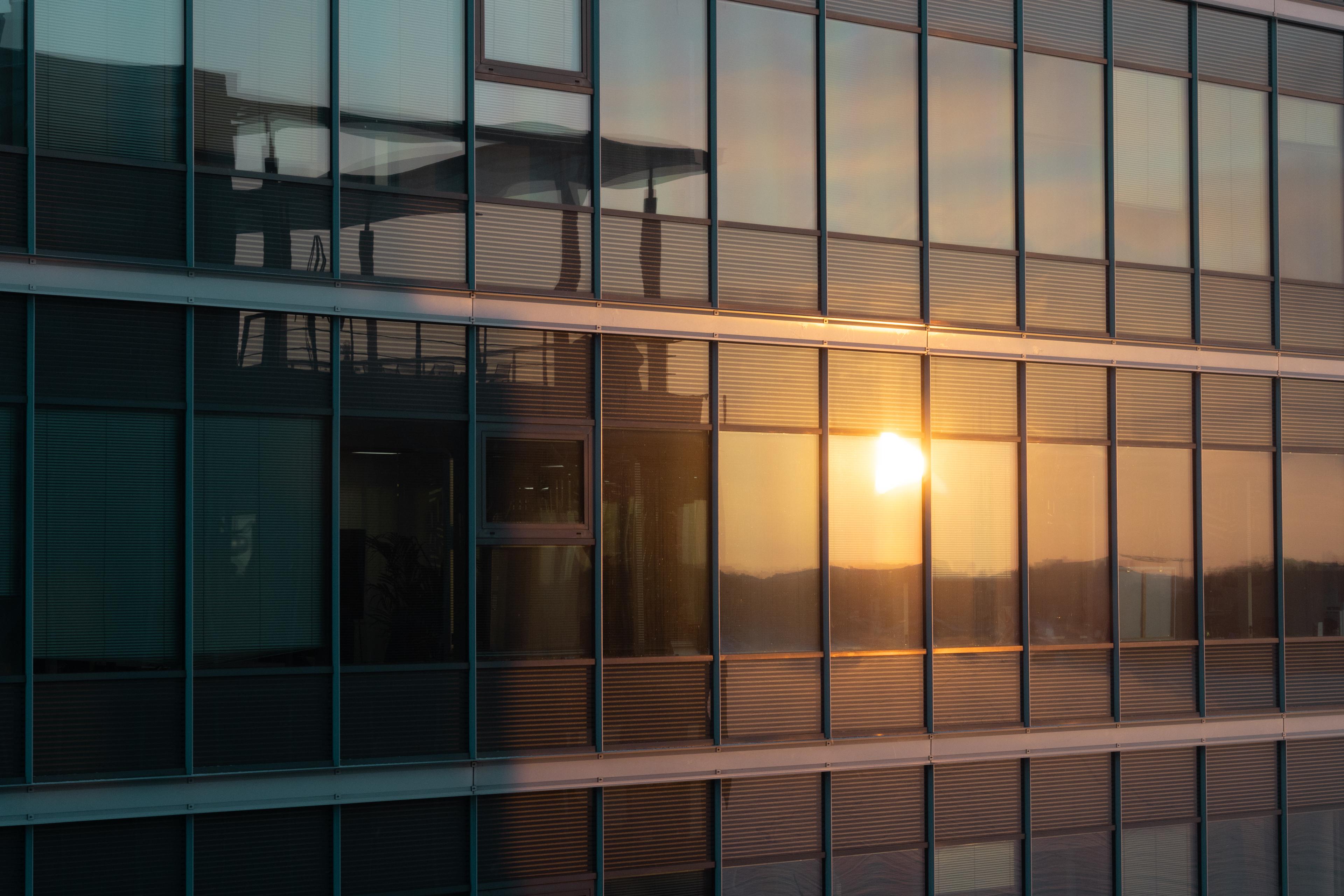 Sun reflecting on a modern glass building's facade at sunset, casting warm light and creating a dynamic interplay of reflections.