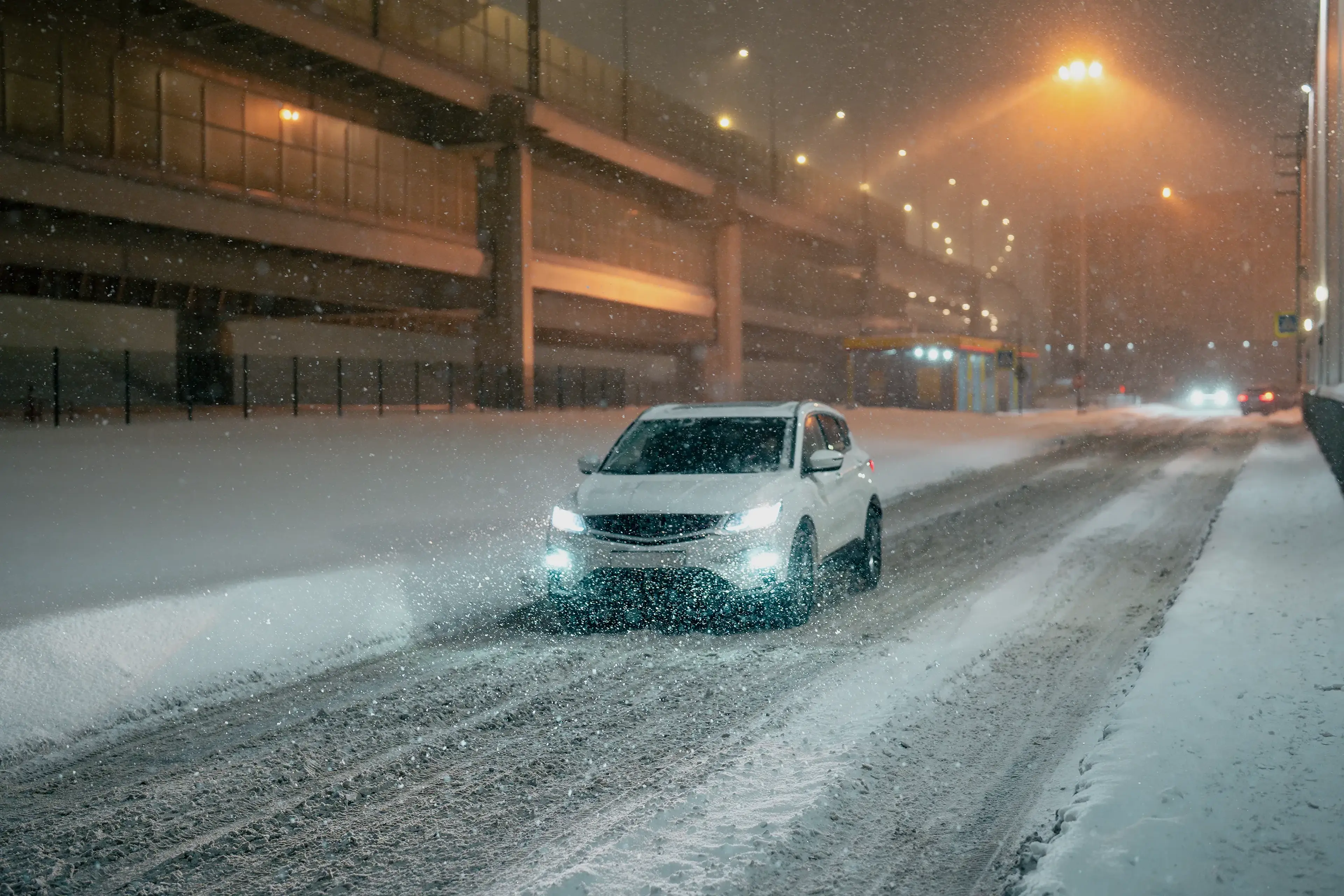 Car driving on a snowy, illuminated urban road at night, with snowflakes falling and streetlights glowing softly.