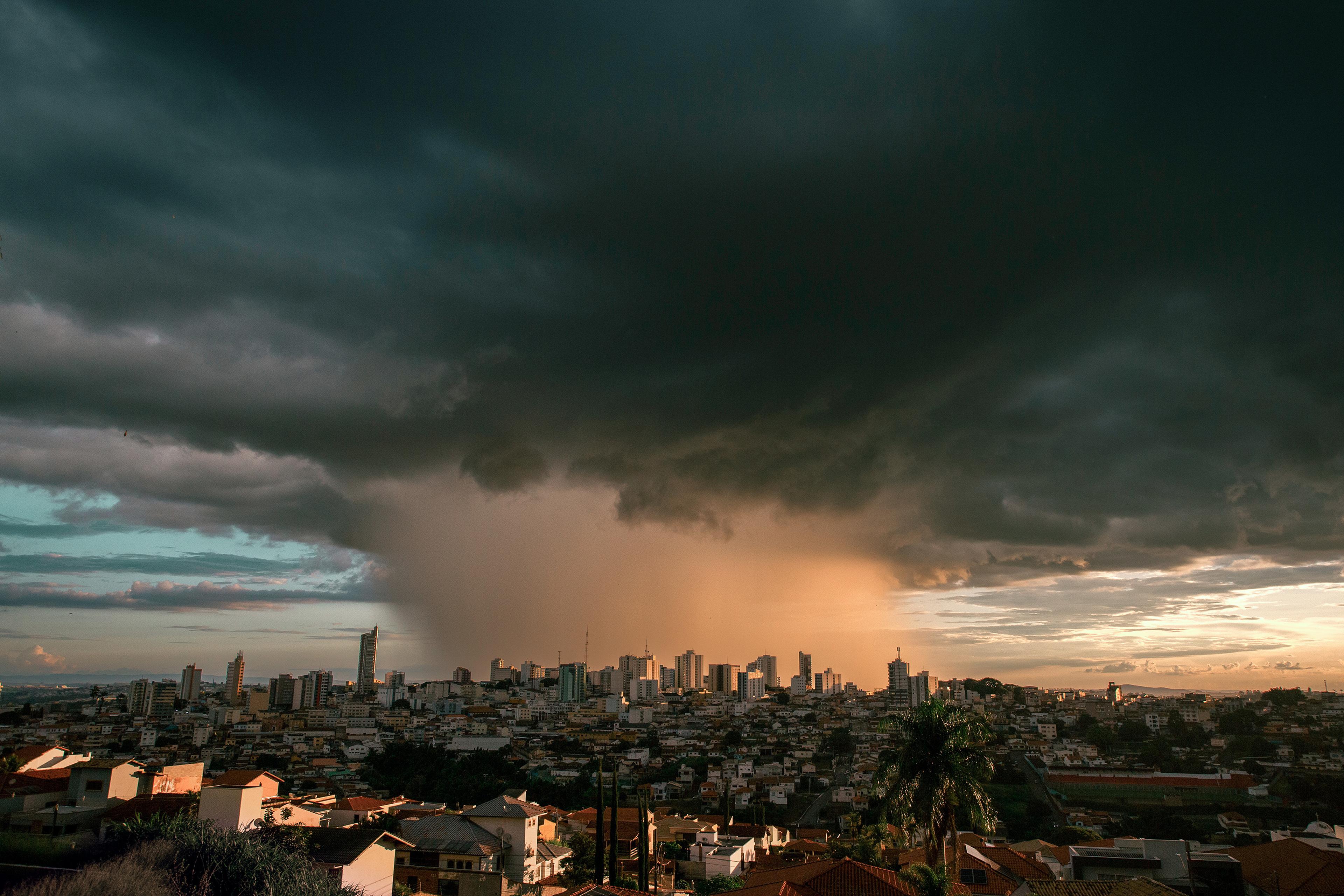 Dark storm clouds loom over a cityscape with buildings silhouetted against an orange sunset, creating a dramatic sky.