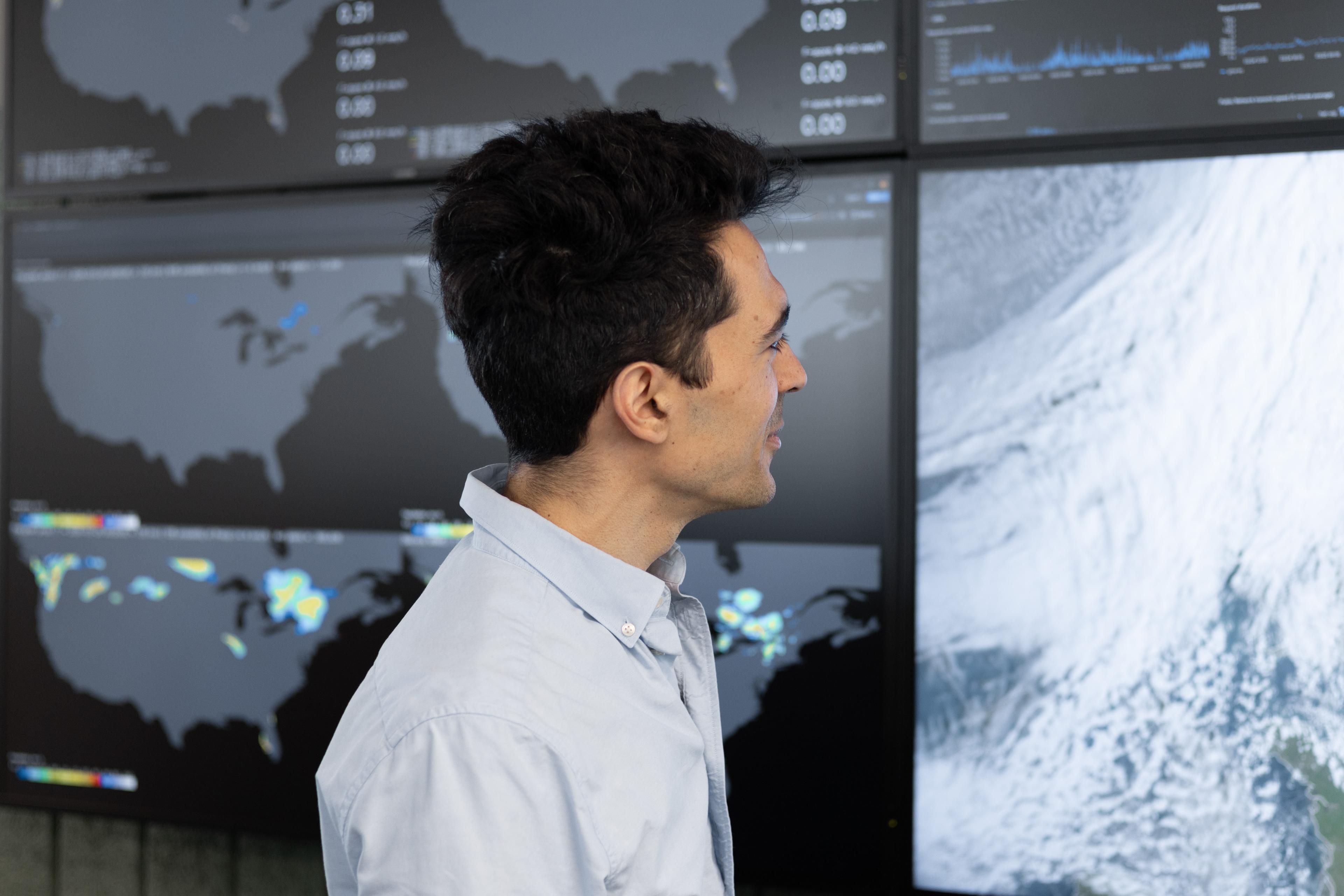 A person in a light blue shirt looks at weather maps displayed on large monitors in a control room setting.
