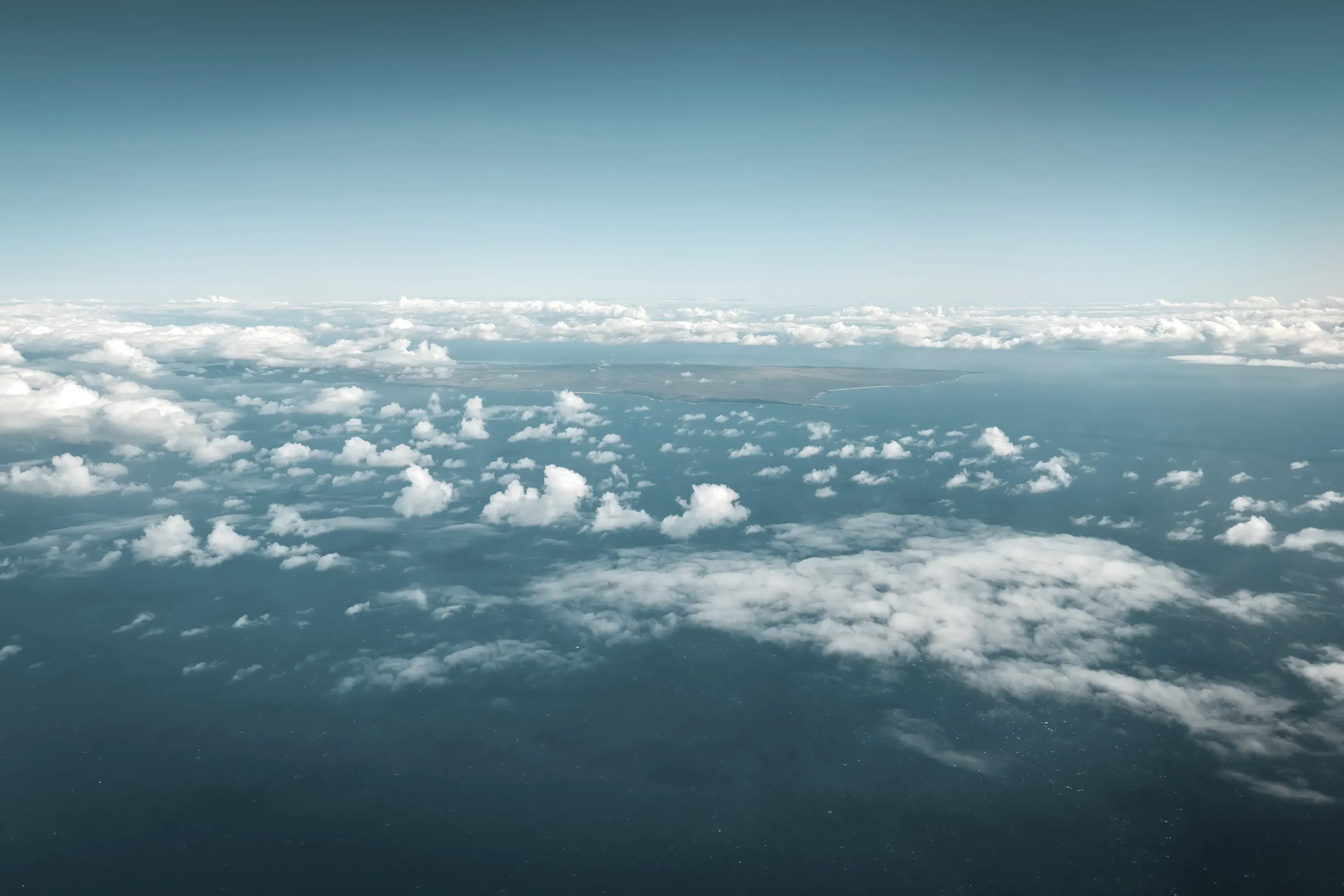 Aerial view of fluffy white clouds over a vast blue ocean, with a faint coastline visible in the distance under a clear sky.