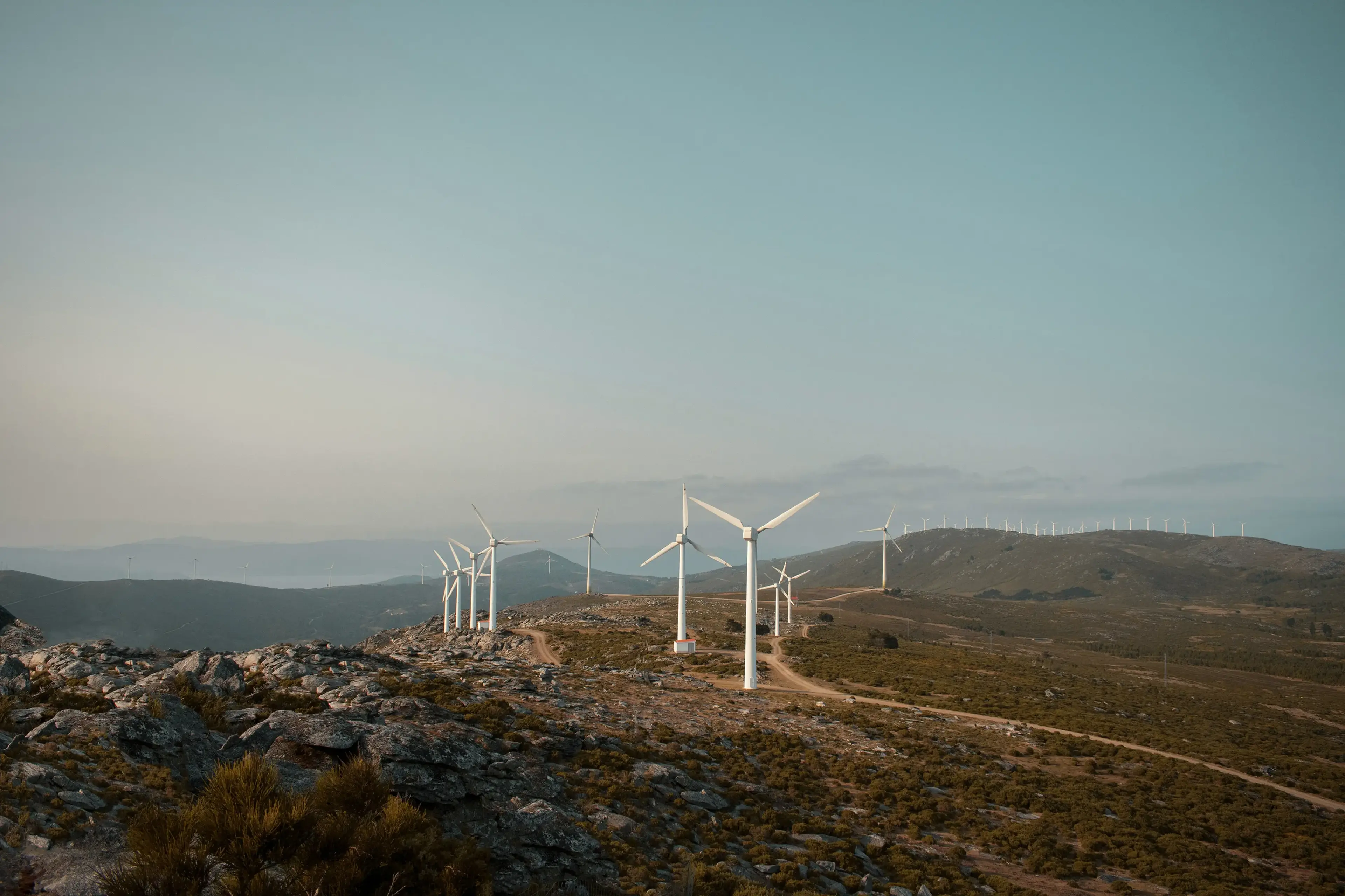 A landscape with several wind turbines on a rocky hilltop under a clear sky, with distant hills in the background.