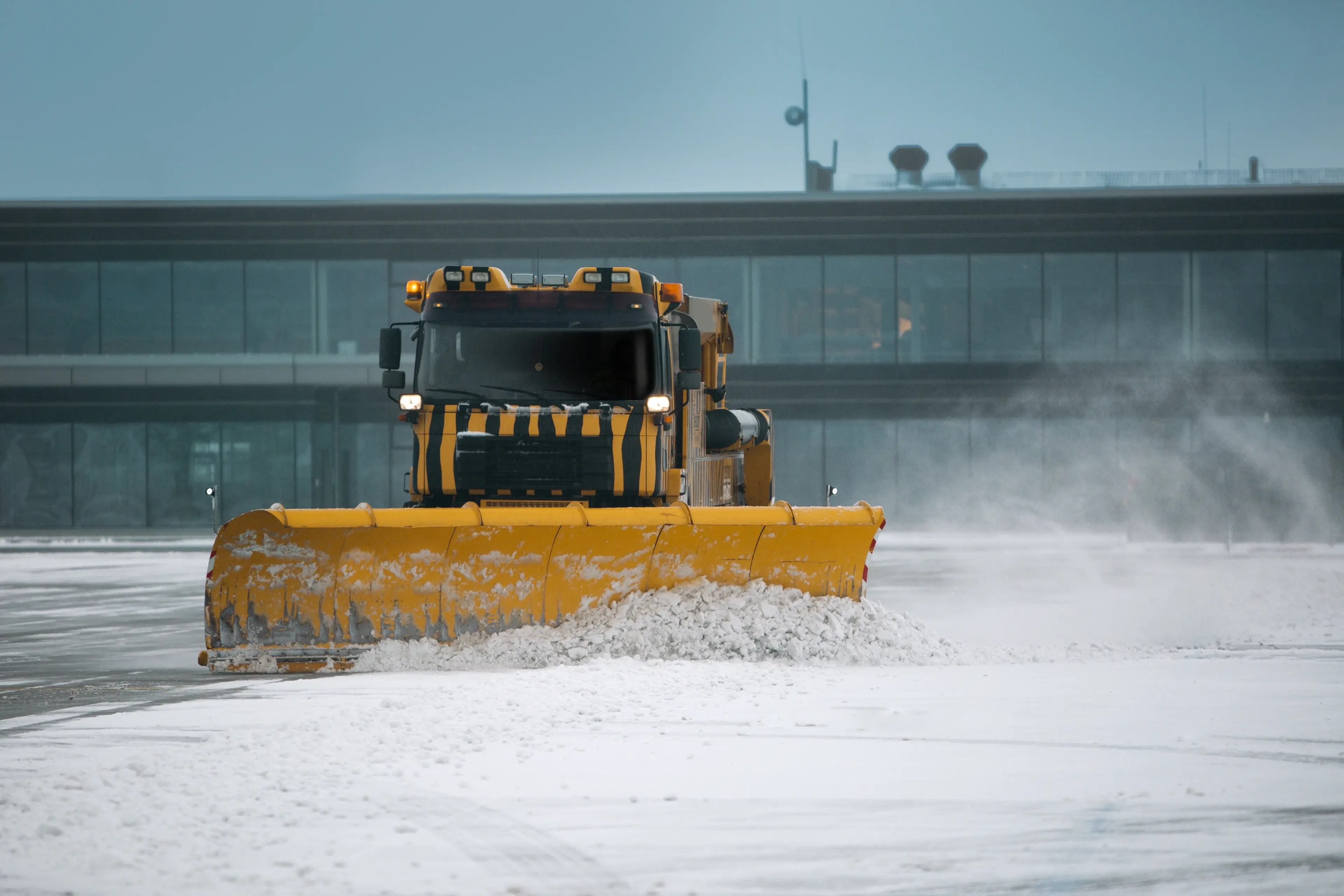A yellow snowplow clearing snow from an airport runway, with a modern terminal building in the background on a cloudy day.