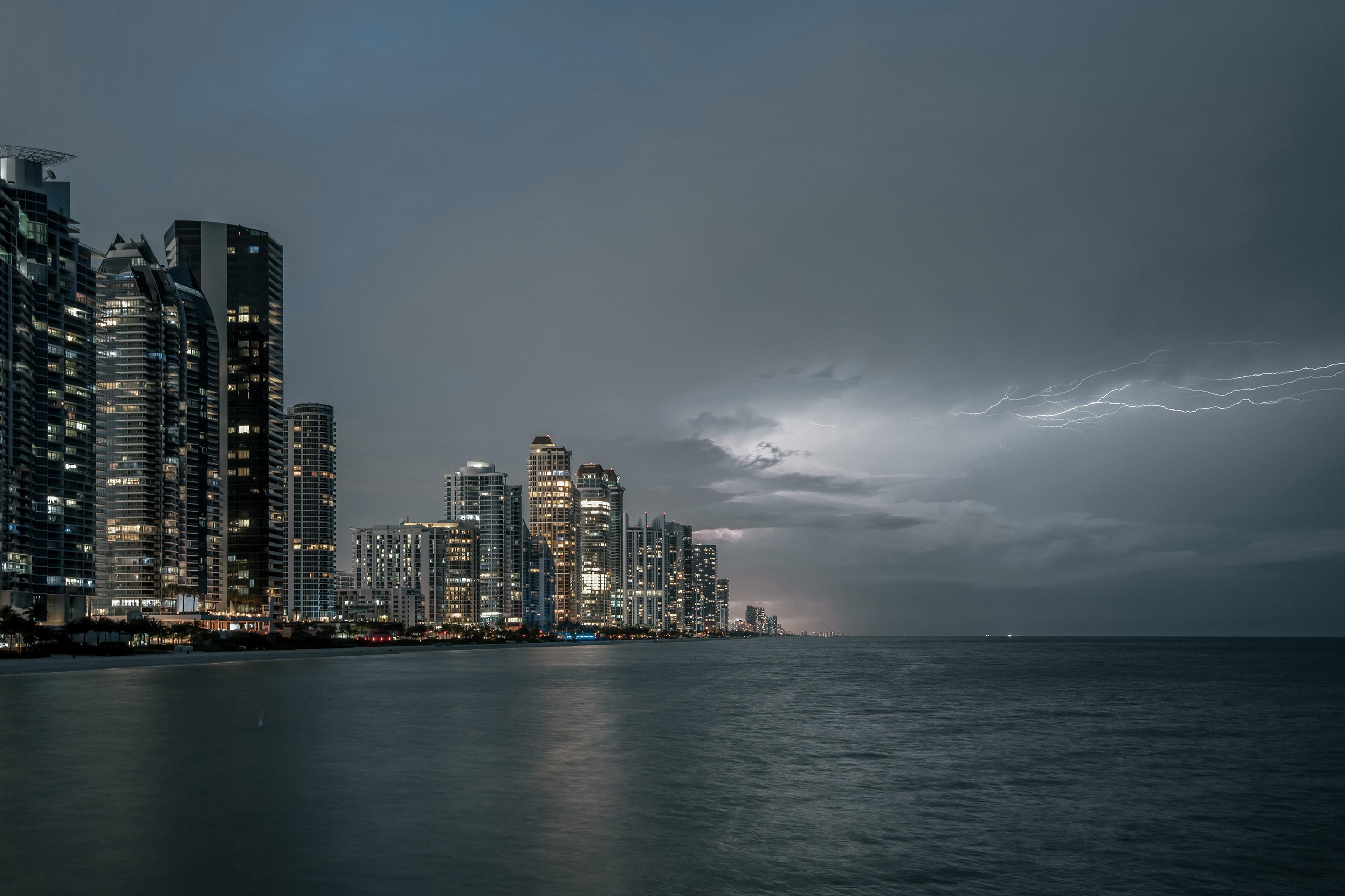 Coastal cityscape at dusk with illuminated skyscrapers and distant lightning flashing over a calm sea under a cloudy sky.