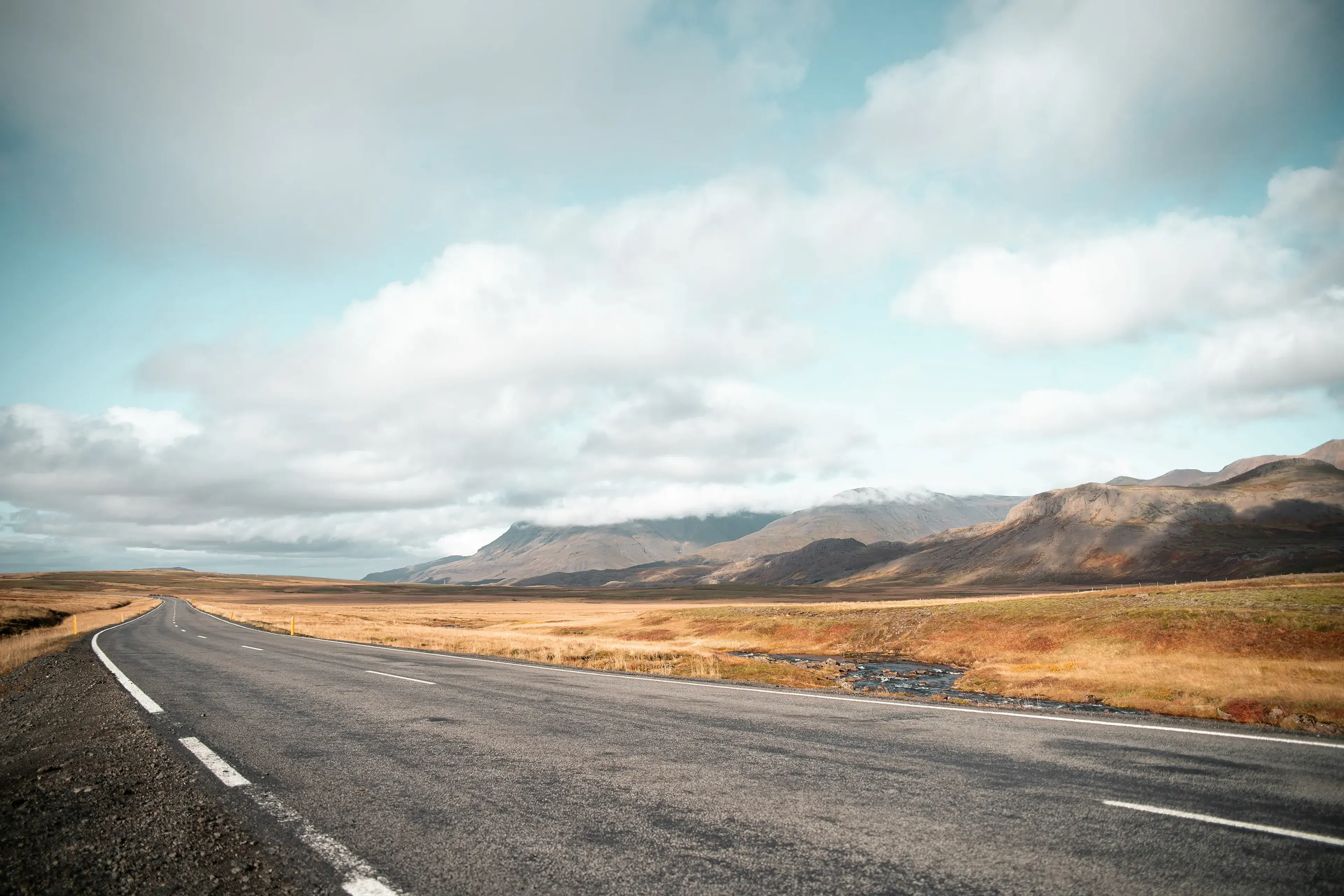 Empty road curving through a vast landscape of golden fields and distant mountains under a blue sky with scattered clouds.