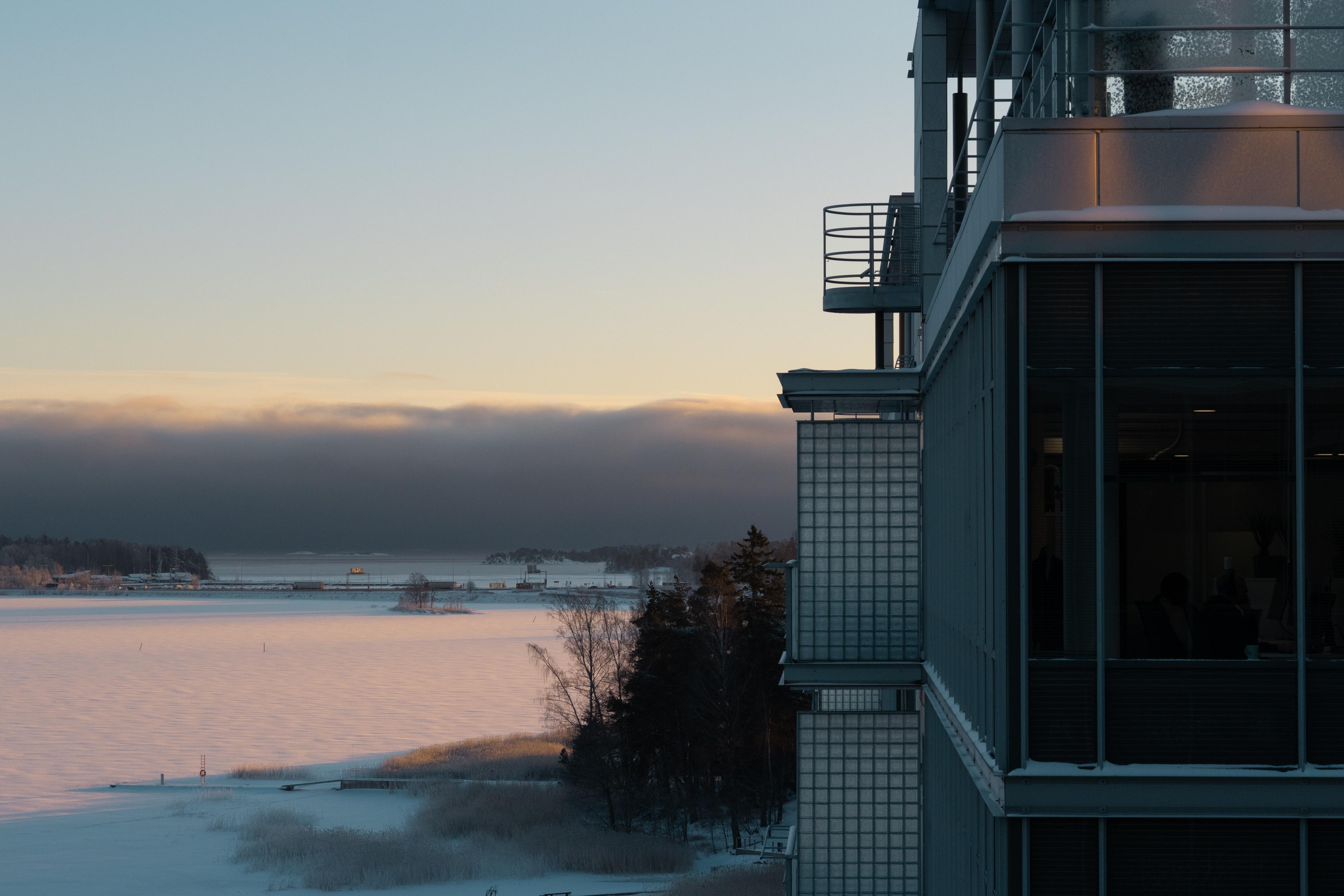 Sunset over a snowy landscape with a modern building on the right. Dark clouds hover above a frozen lake and distant trees.