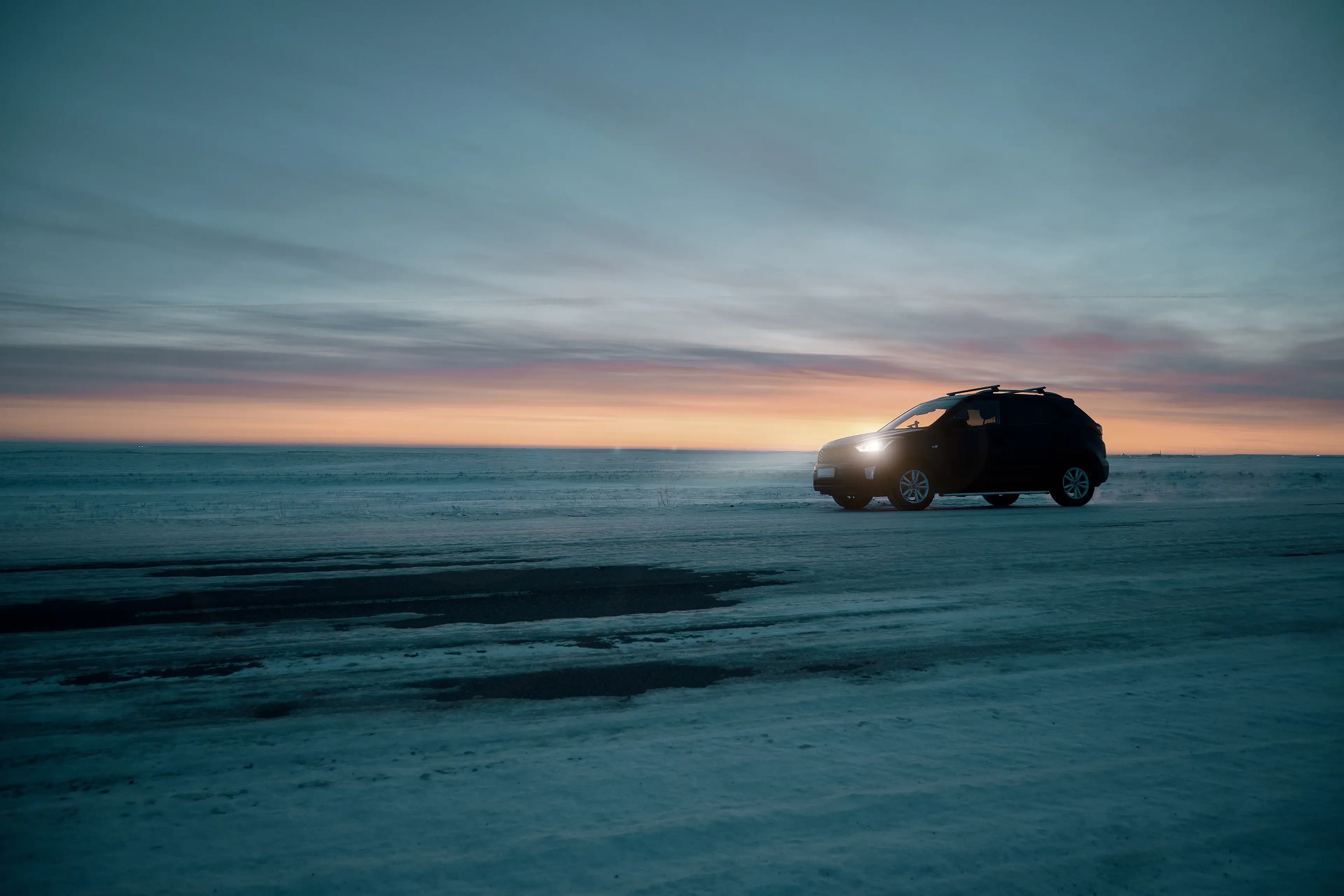 SUV driving on a snowy road at twilight, with a colorful sunset sky in the background.