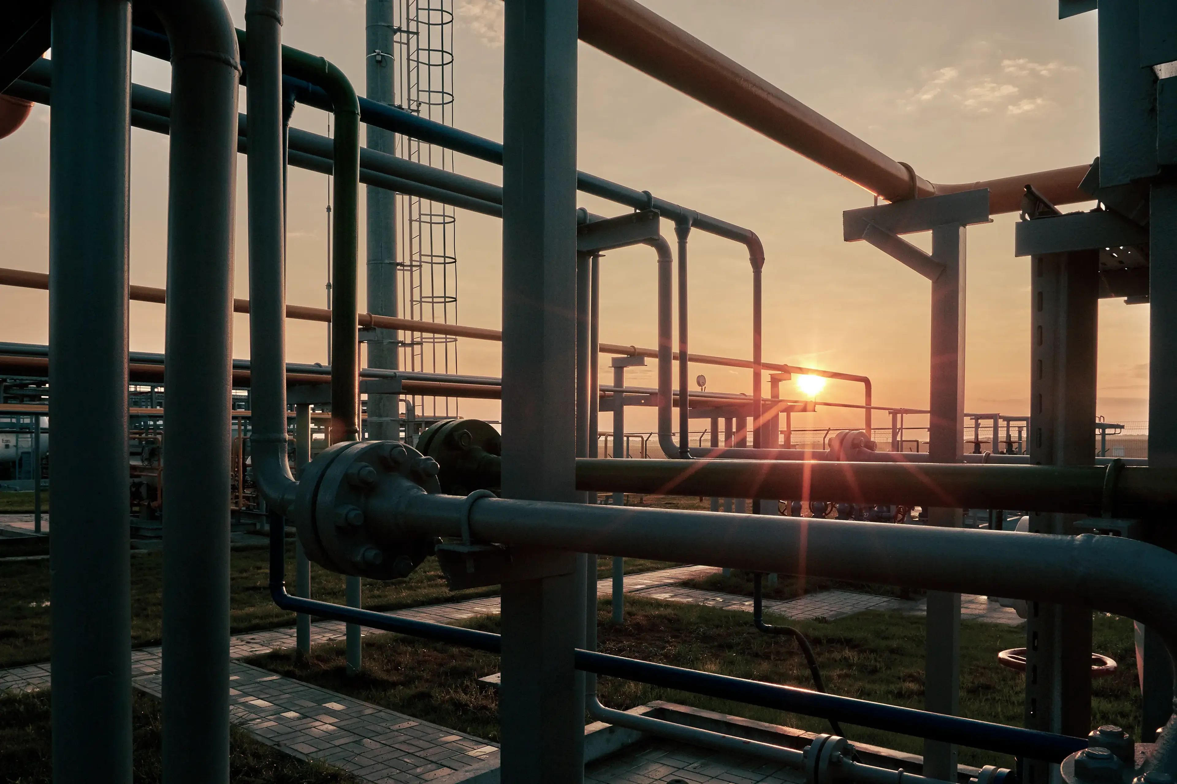 Industrial pipes silhouetted against a sunset, with orange and blue lighting, creating a dramatic contrast and intricate pattern.
