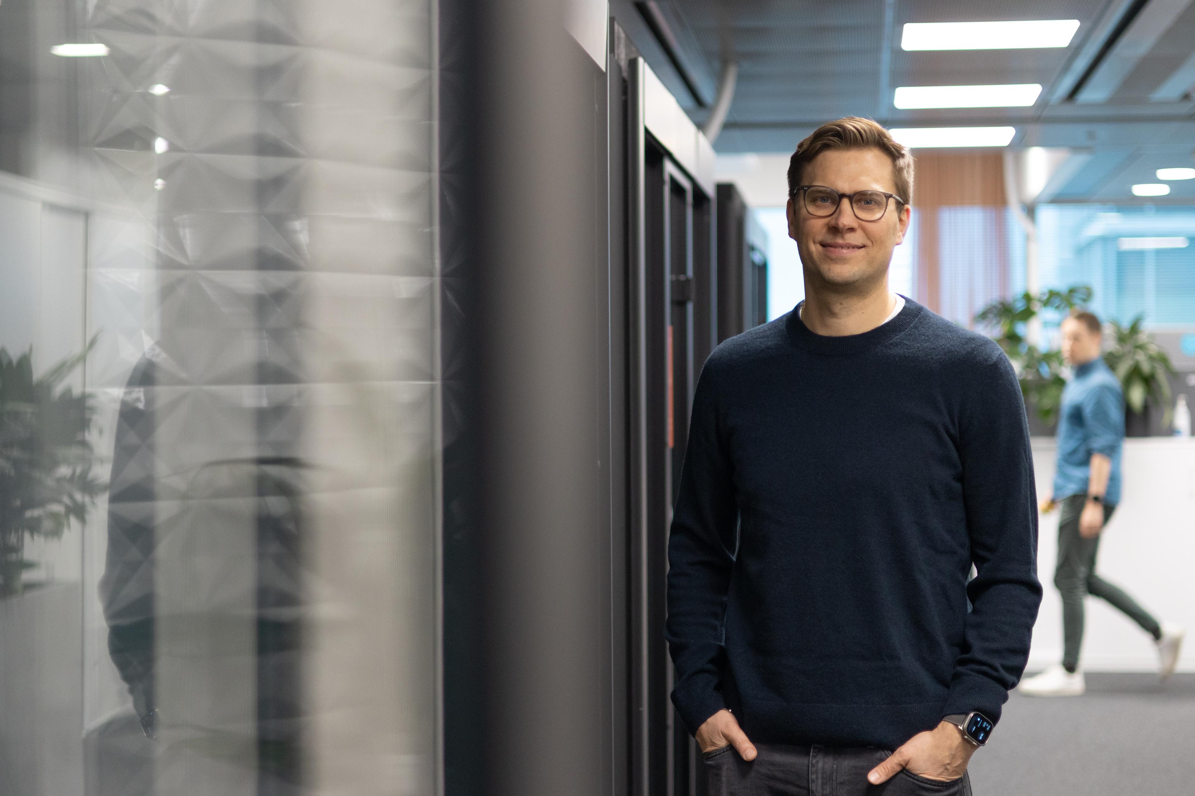 A smiling man with glasses stands in an office hallway, hands in pockets. Another person walks in the background.