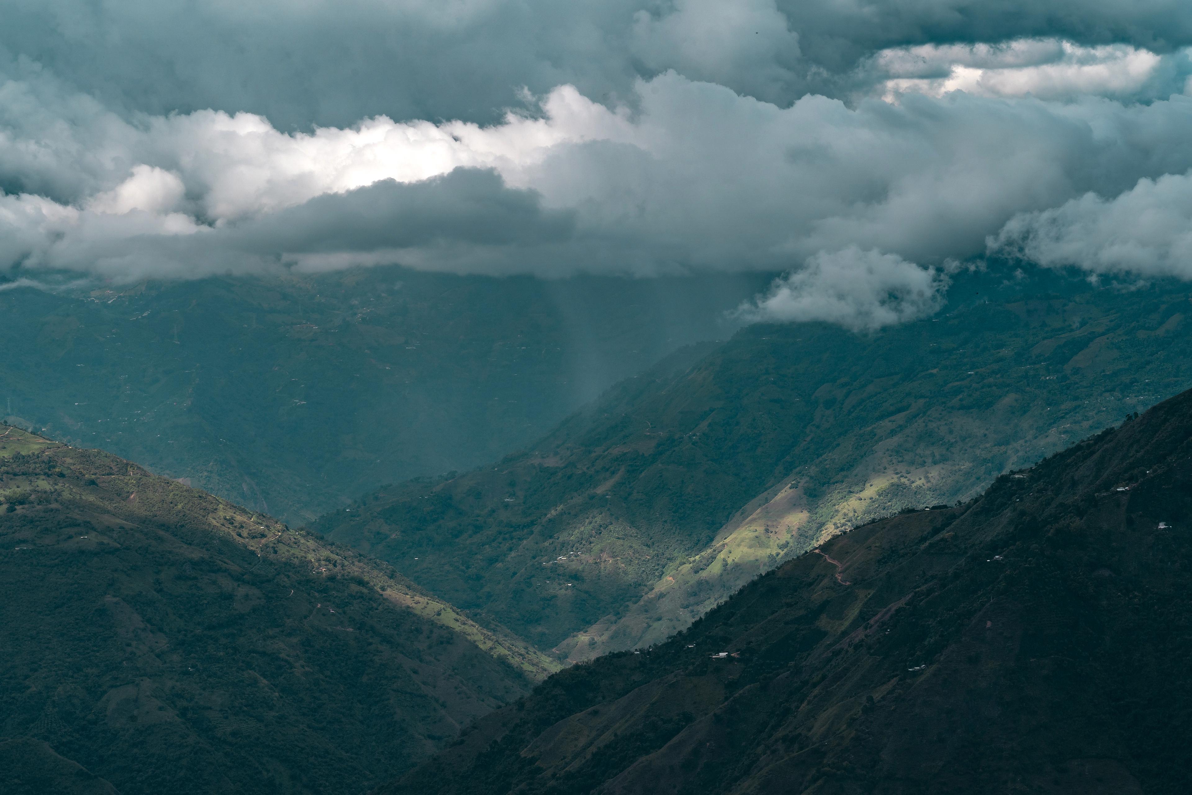A scenic mountain valley under a cloudy sky, with patches of sunlight highlighting the lush green slopes and forests below.