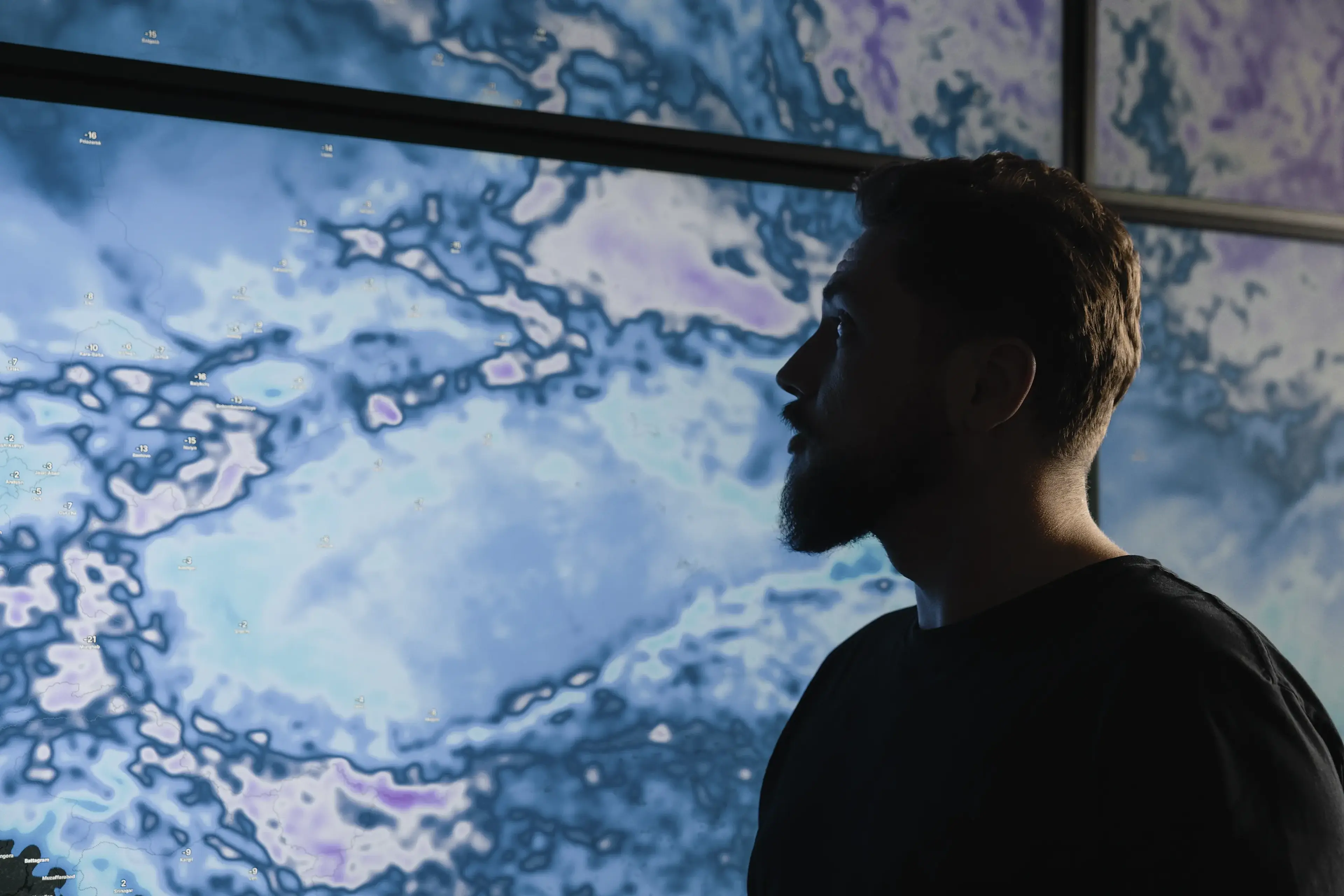 A man in silhouette looks at a large display with blue and purple weather map patterns