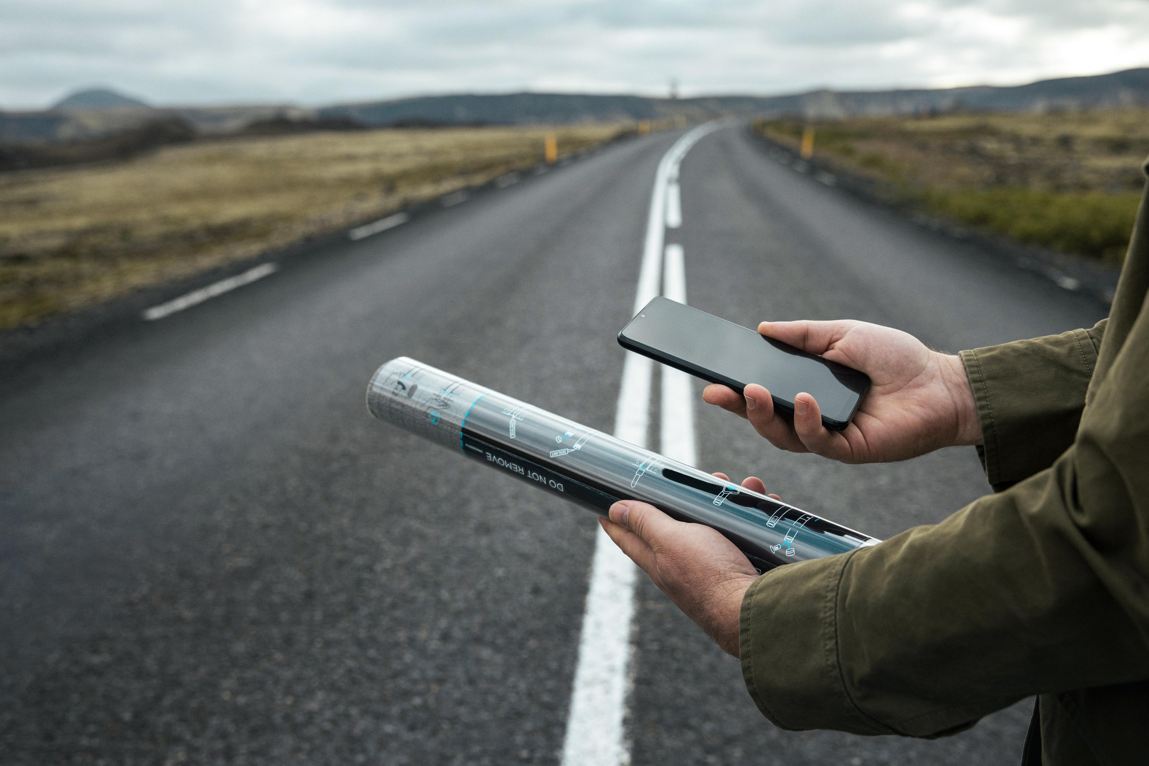 A person holds a smartphone and a Vaisala GroundCast sensor while standing on an empty road under a cloudy sky.