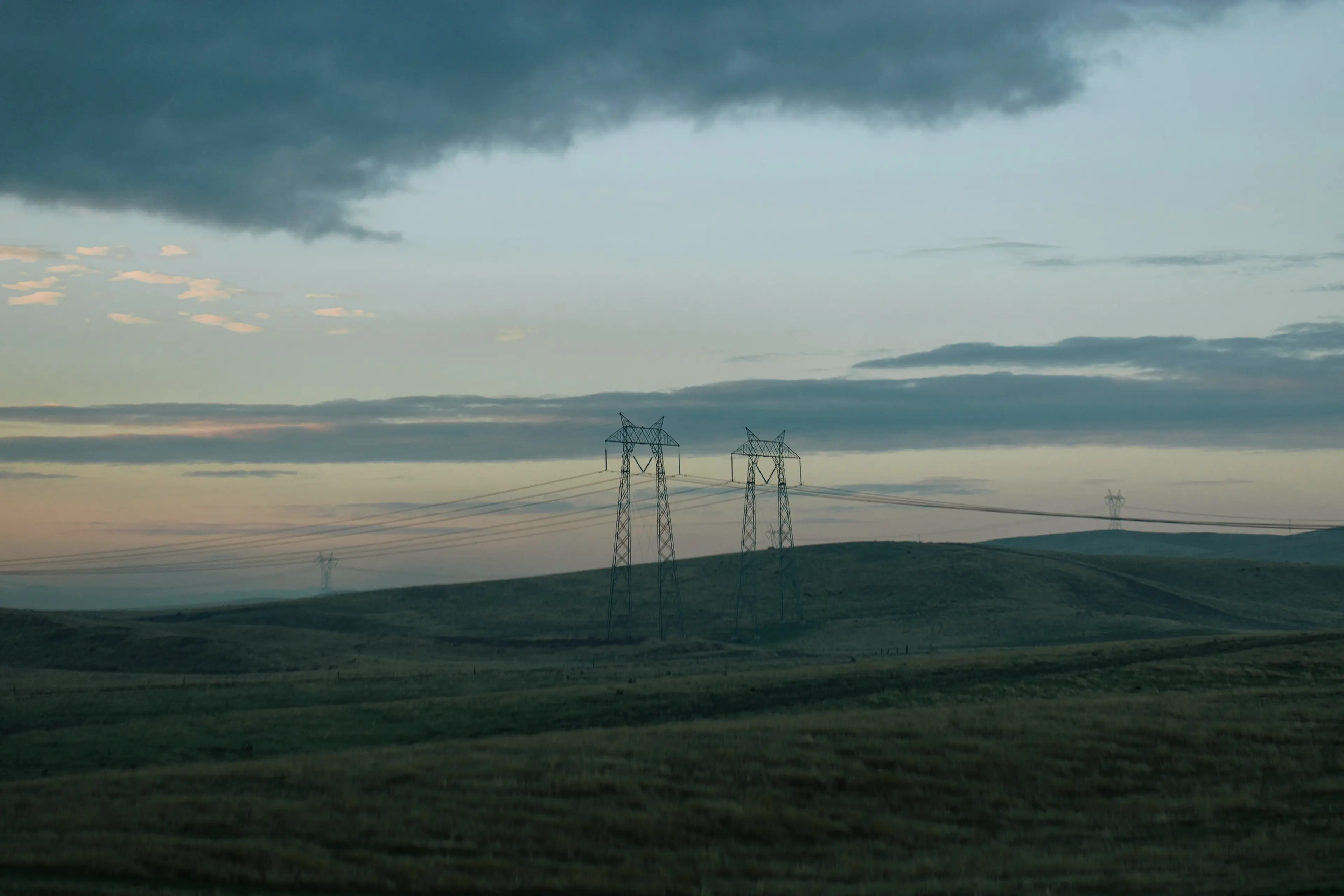 Silhouetted power lines stretch across a grassy rolling landscape under a cloudy, dusk sky.