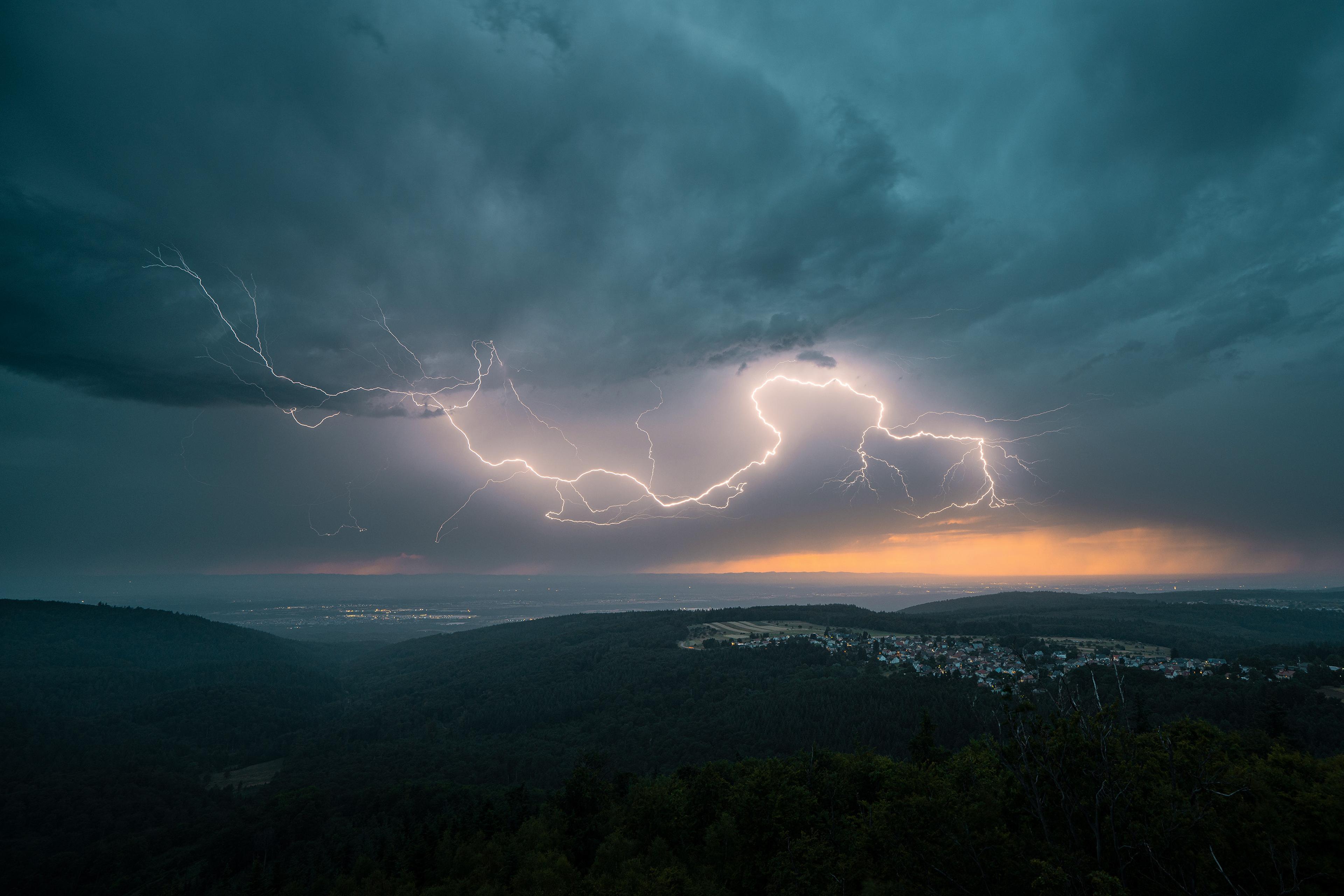A dramatic landscape with bright lightning streaks across a dark, cloudy sky above a forest and small town during sunset.