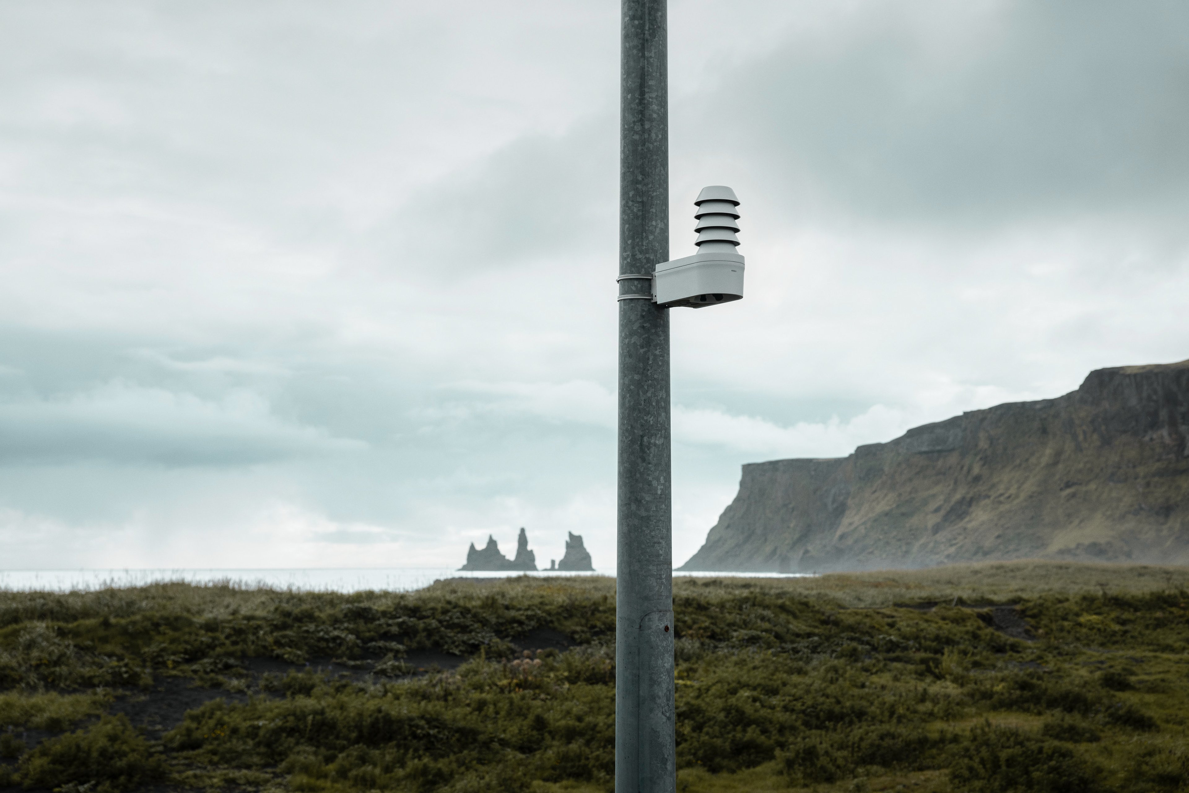 A Vaisala Xweather TempCast weather sensor mounted on a pole in a grassy area with rocky sea stacks and cliffs in the cloudy background.
