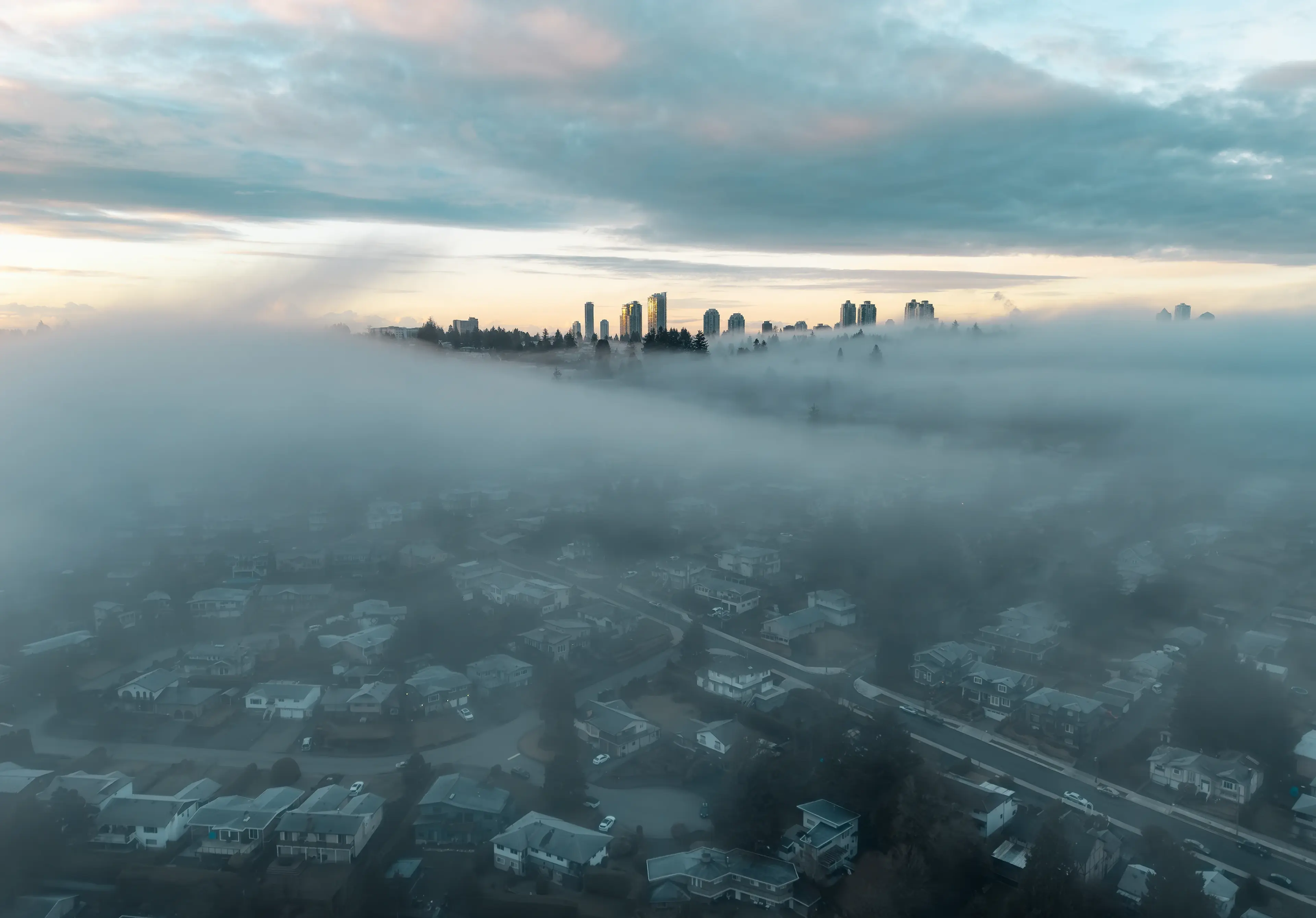 Foggy cityscape with high-rise buildings emerging above dense fog, blanketing suburban houses below, under a moody sky at sunset.