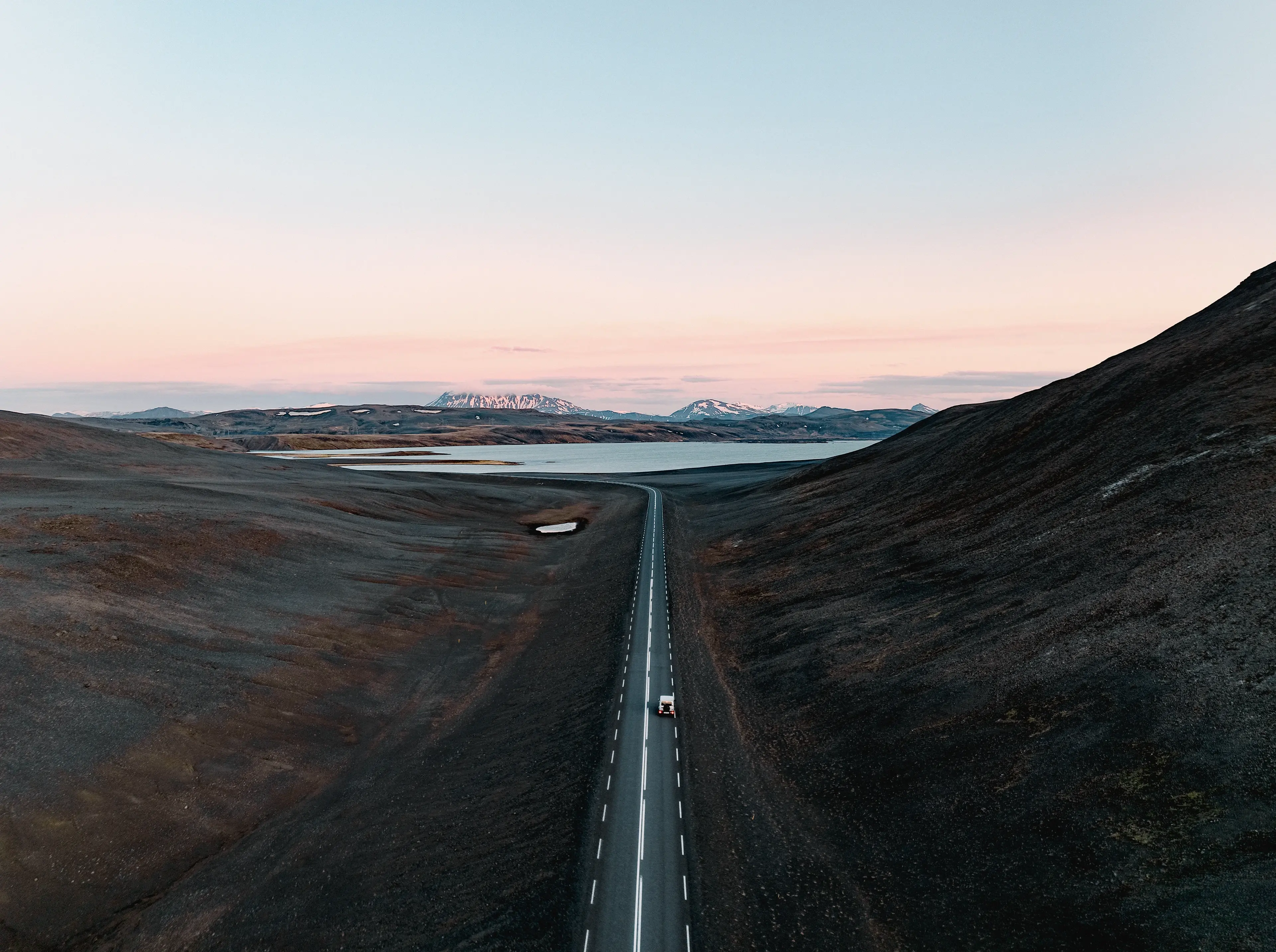 A solitary car drives on a long, straight road through a barren landscape with hills, a lake, and snow-capped mountains under a pastel sky.
