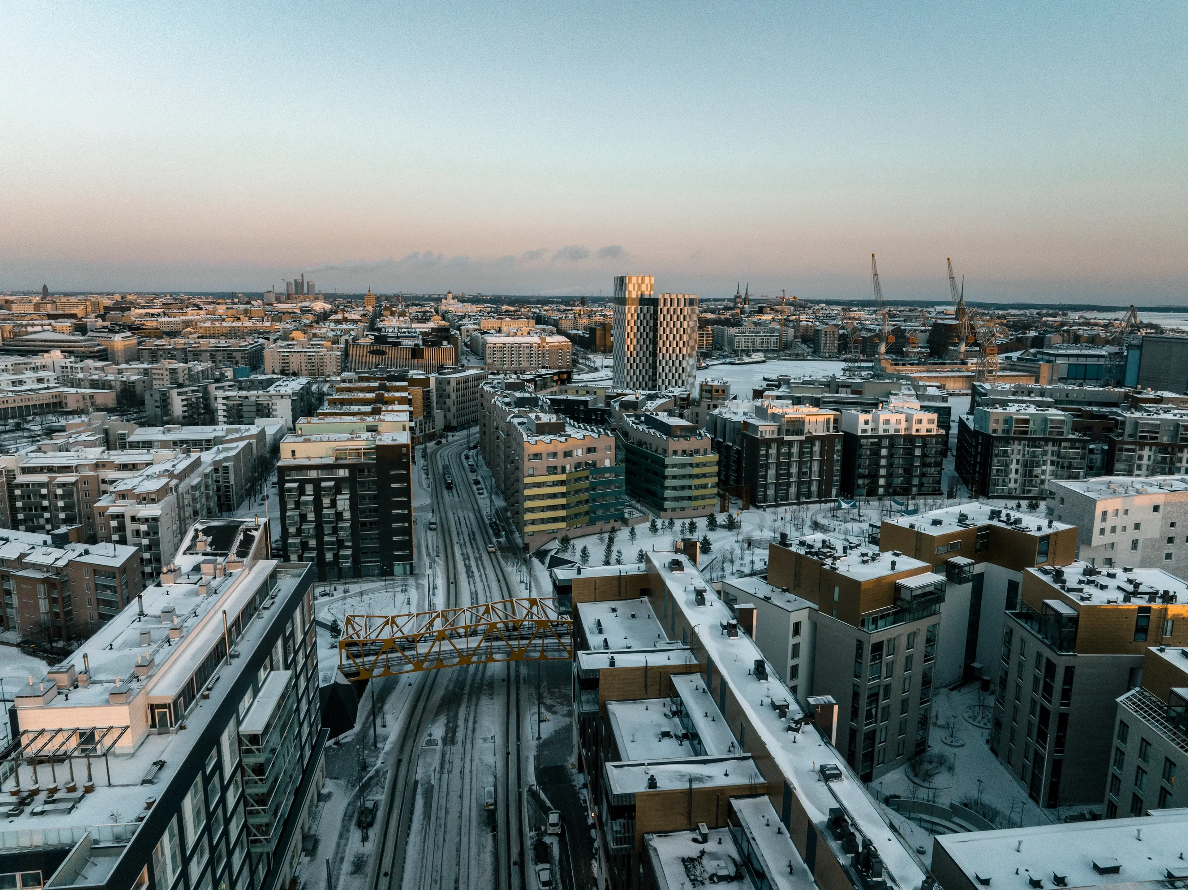 Aerial view of a snowy urban landscape at sunset, featuring modern buildings and a bridge over a road.