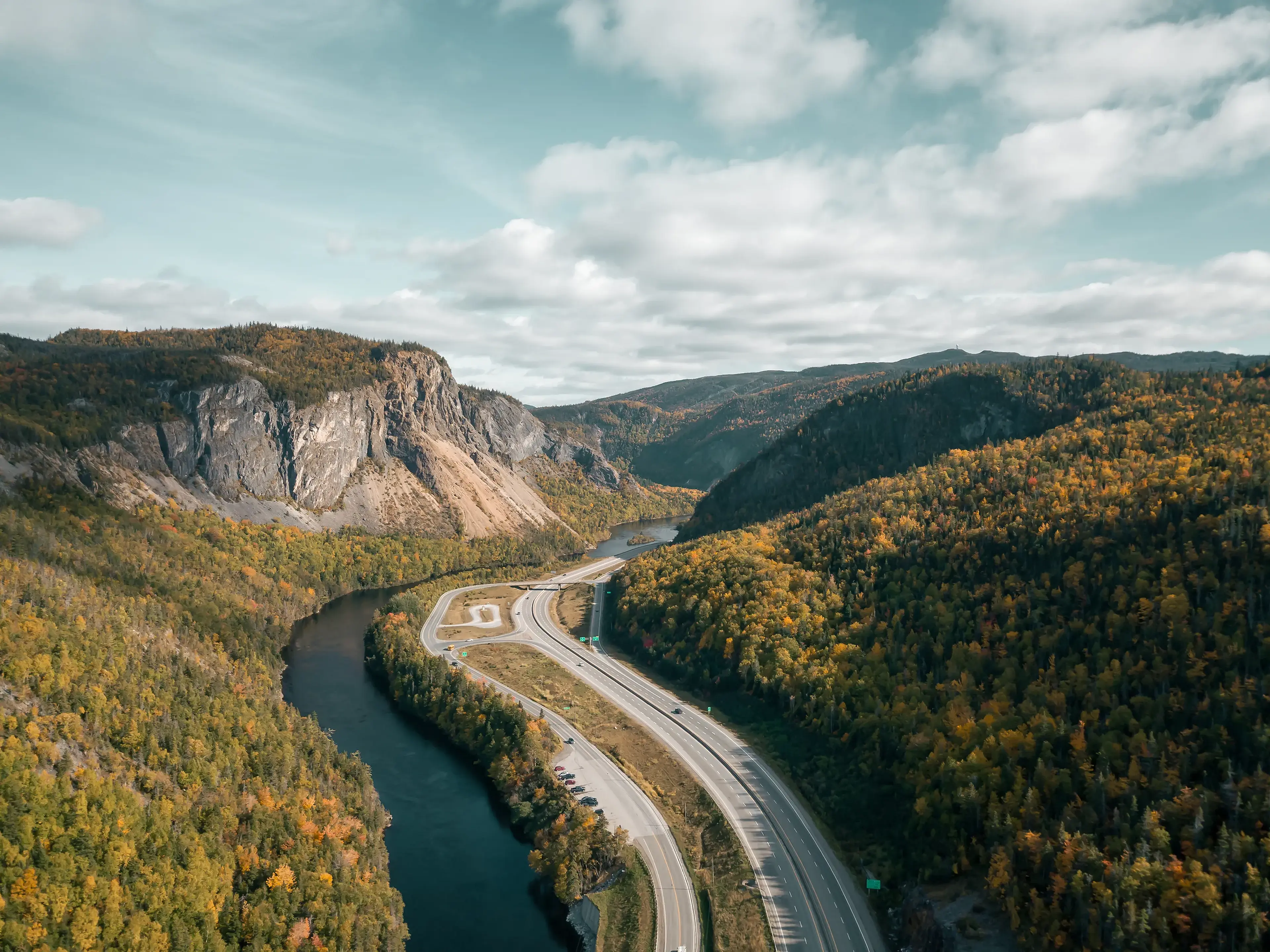 Aerial view of a scenic road winding through a forested valley with a river, surrounded by rugged mountains under a partly cloudy sky.