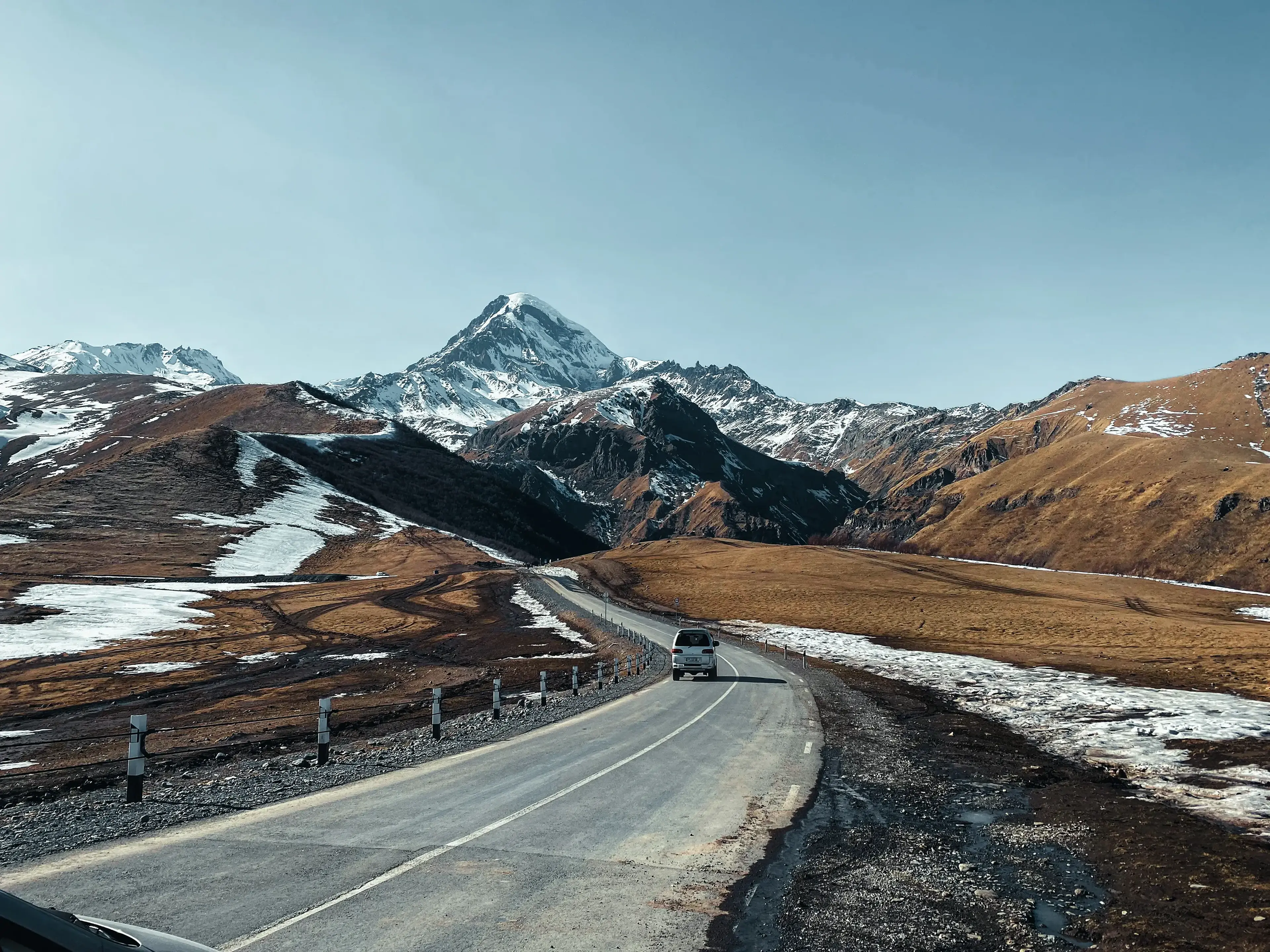 A vehicle travels on a paved road through a mountainous landscape with patches of snow and a prominent peak under a clear blue sky.