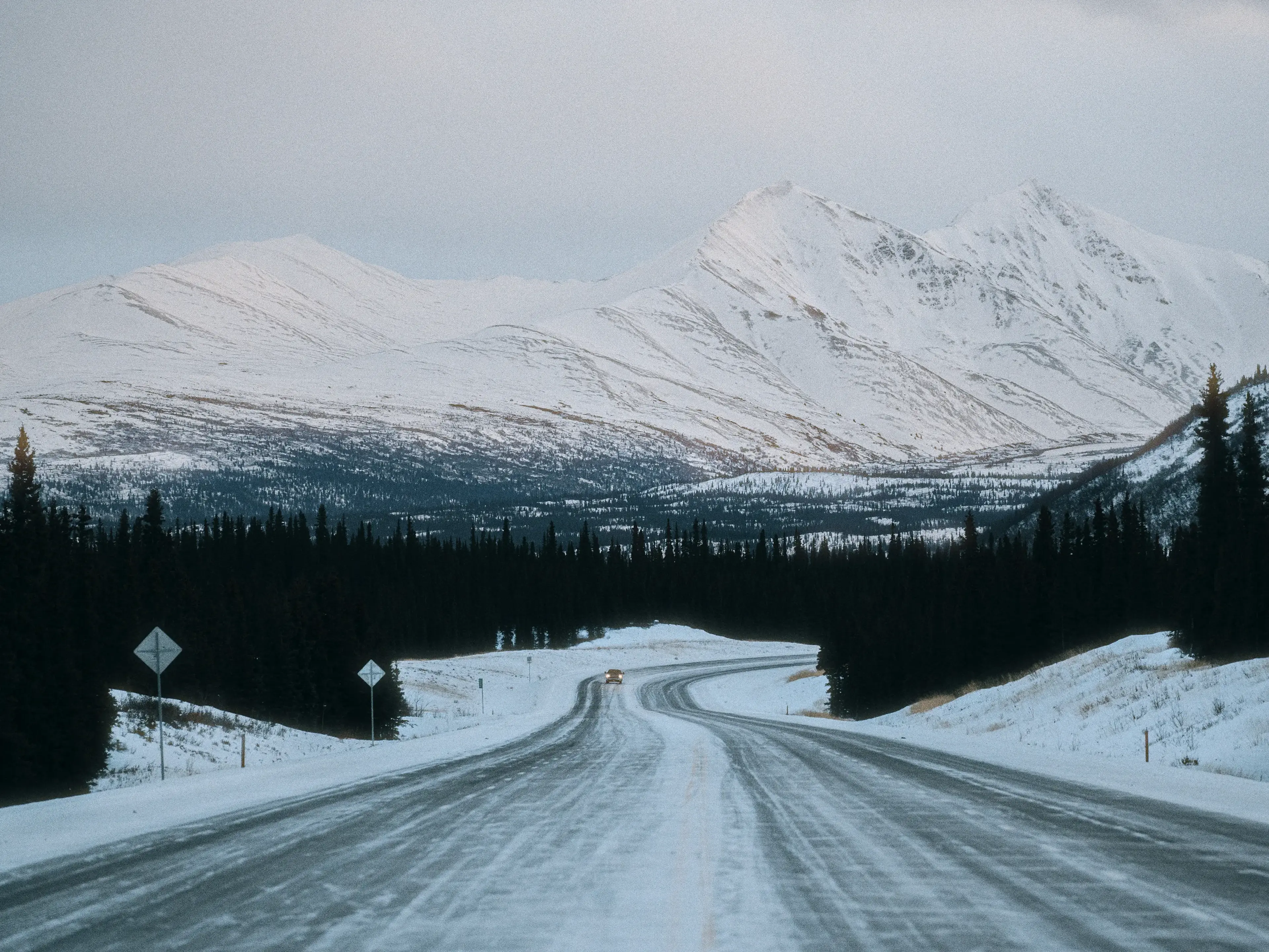 Snow-covered road leading to distant mountains under a cloudy sky, flanked by evergreen trees, with a car approaching in the distance.