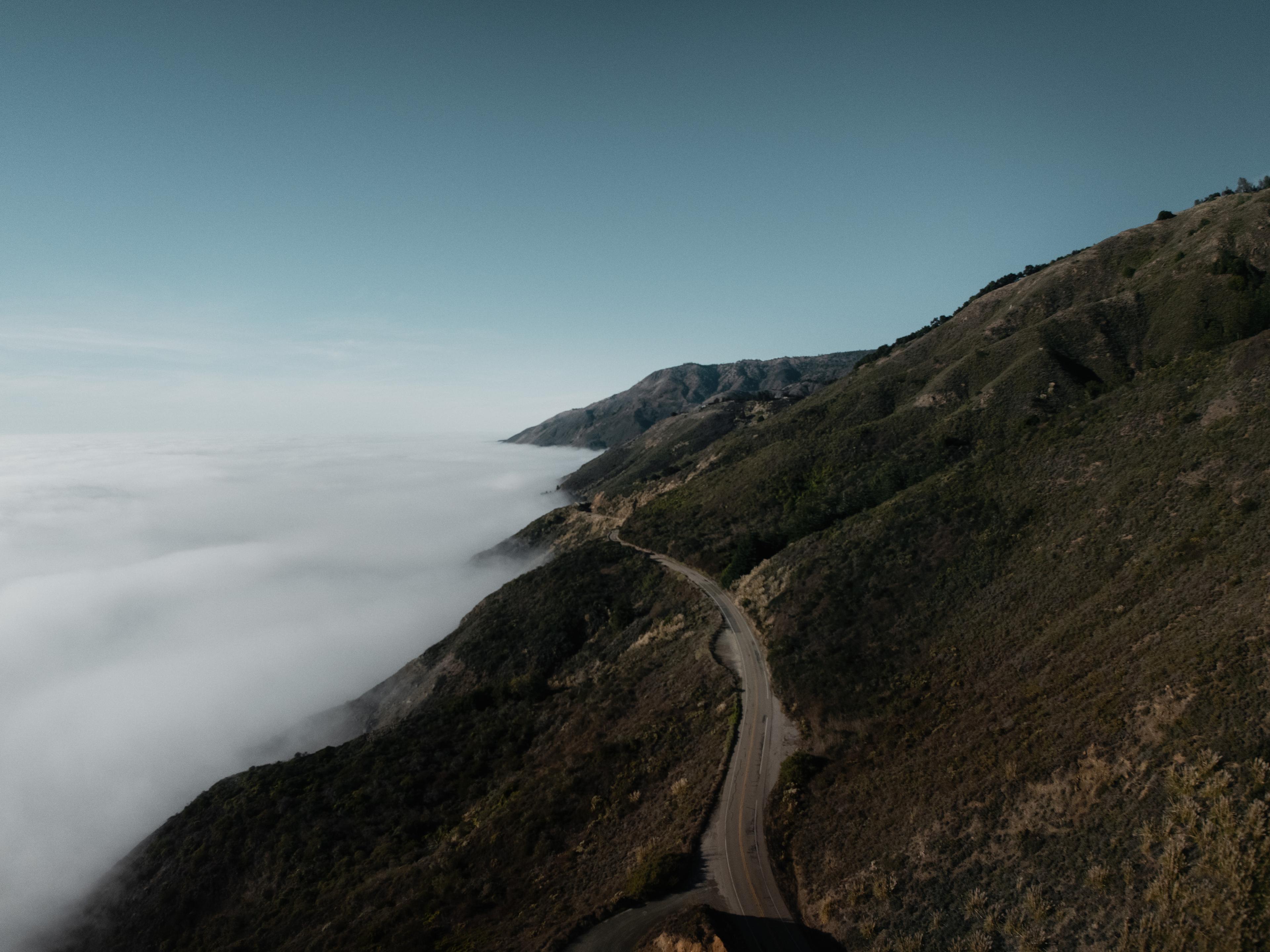 A winding road curves along a lush, mountainous coast with a blanket of clouds over the ocean under a clear blue sky.