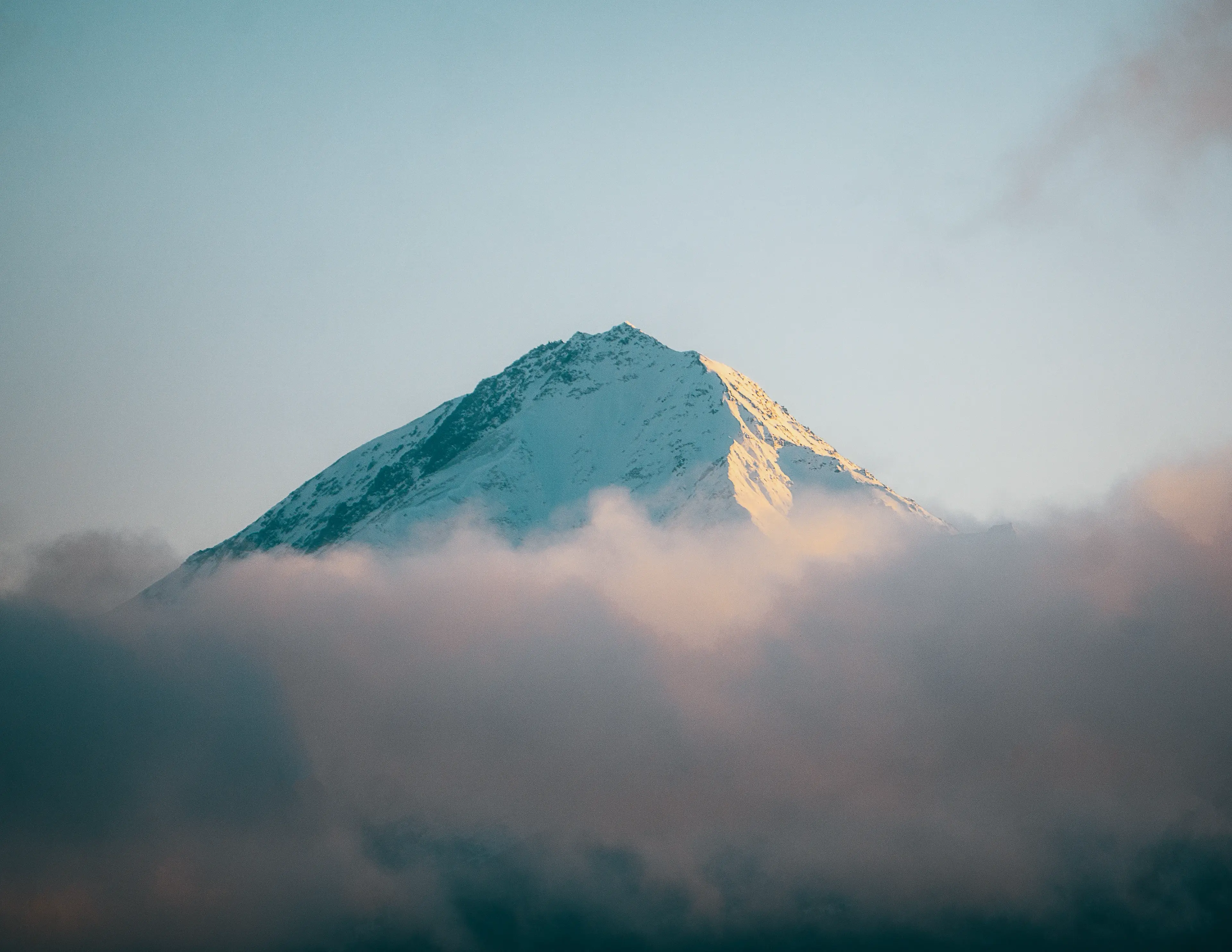 Snow-capped mountain peak partially obscured by soft, pink-tinted clouds against a clear blue sky.