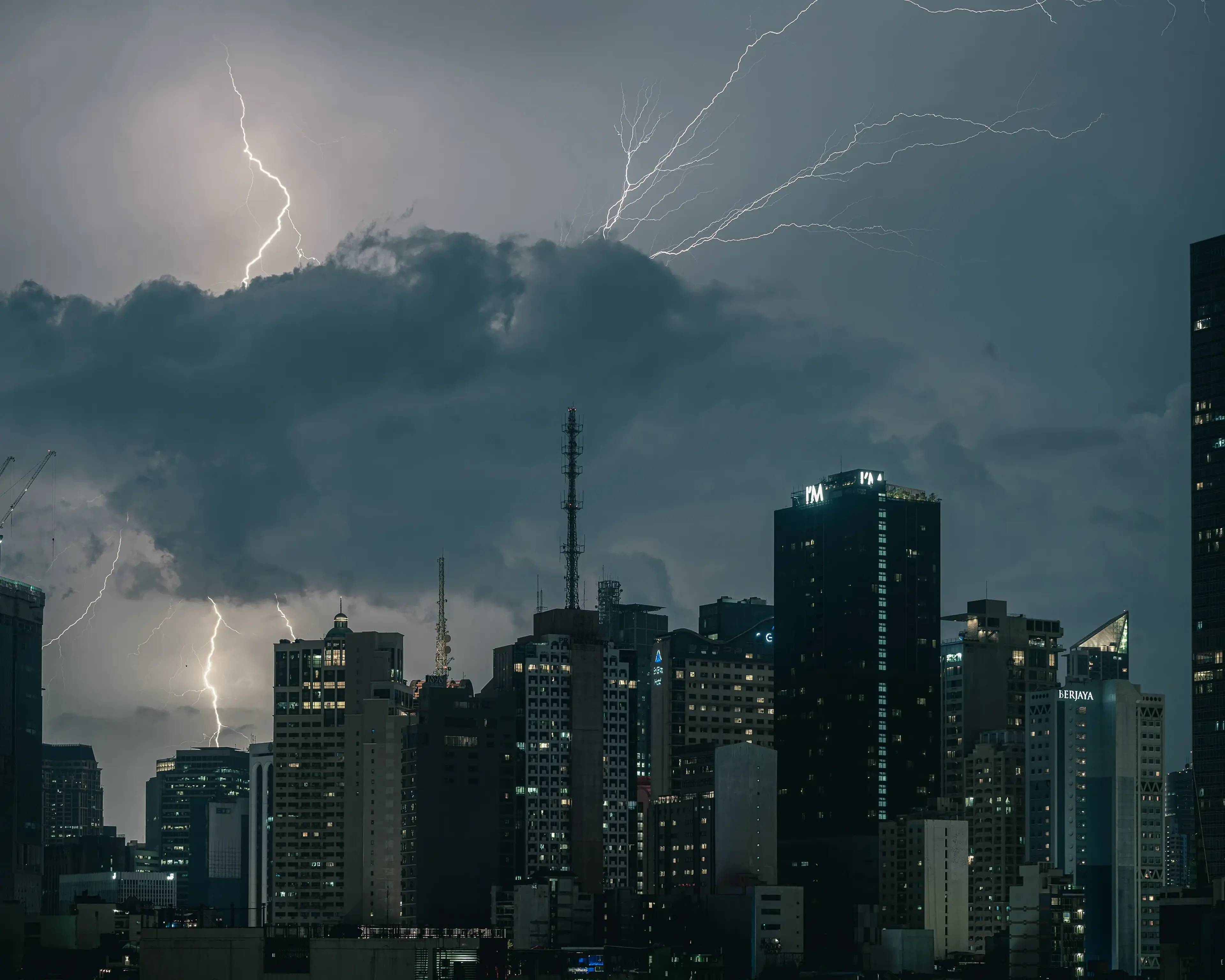 City skyline at night with tall buildings under a dark, cloudy sky. Lightning strikes illuminate the clouds, creating a dramatic scene.