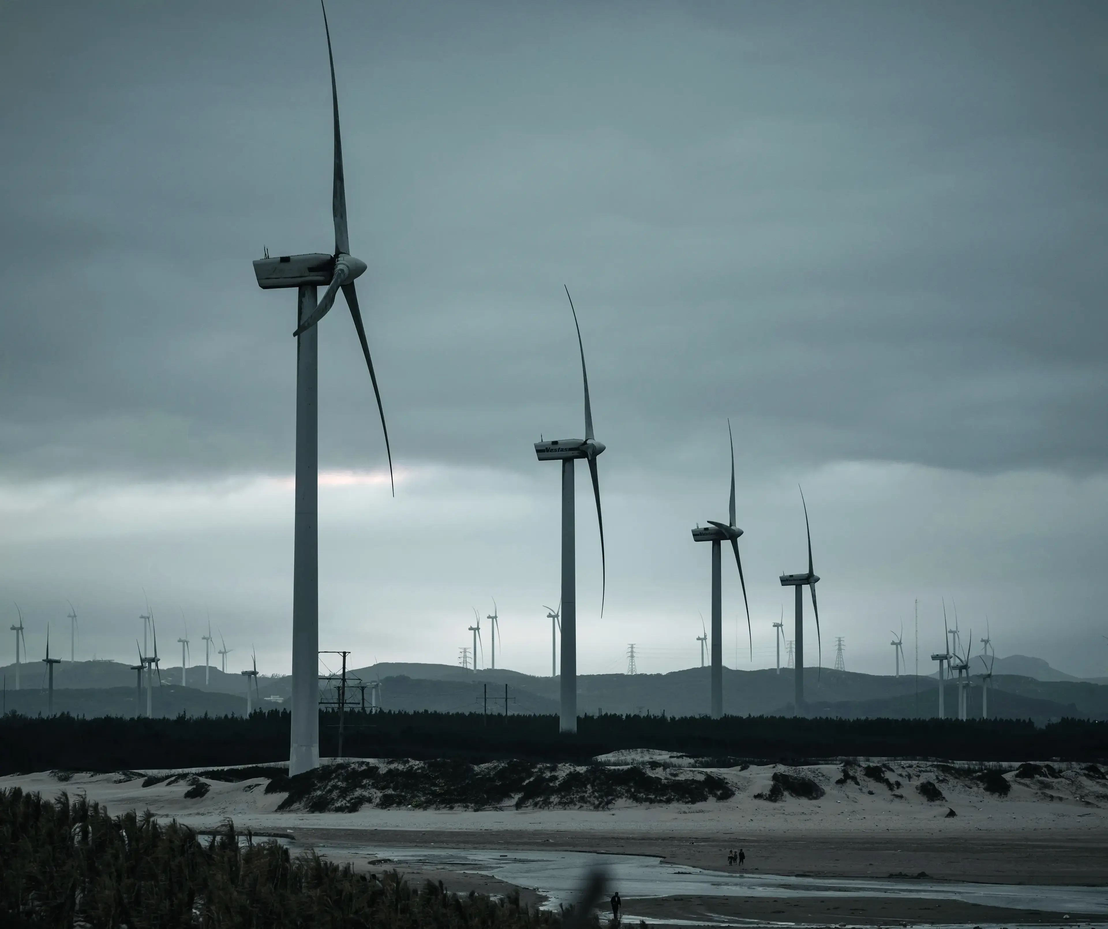 Wind turbines on a cloudy day, set against a distant mountain range and sandy beach in the foreground.