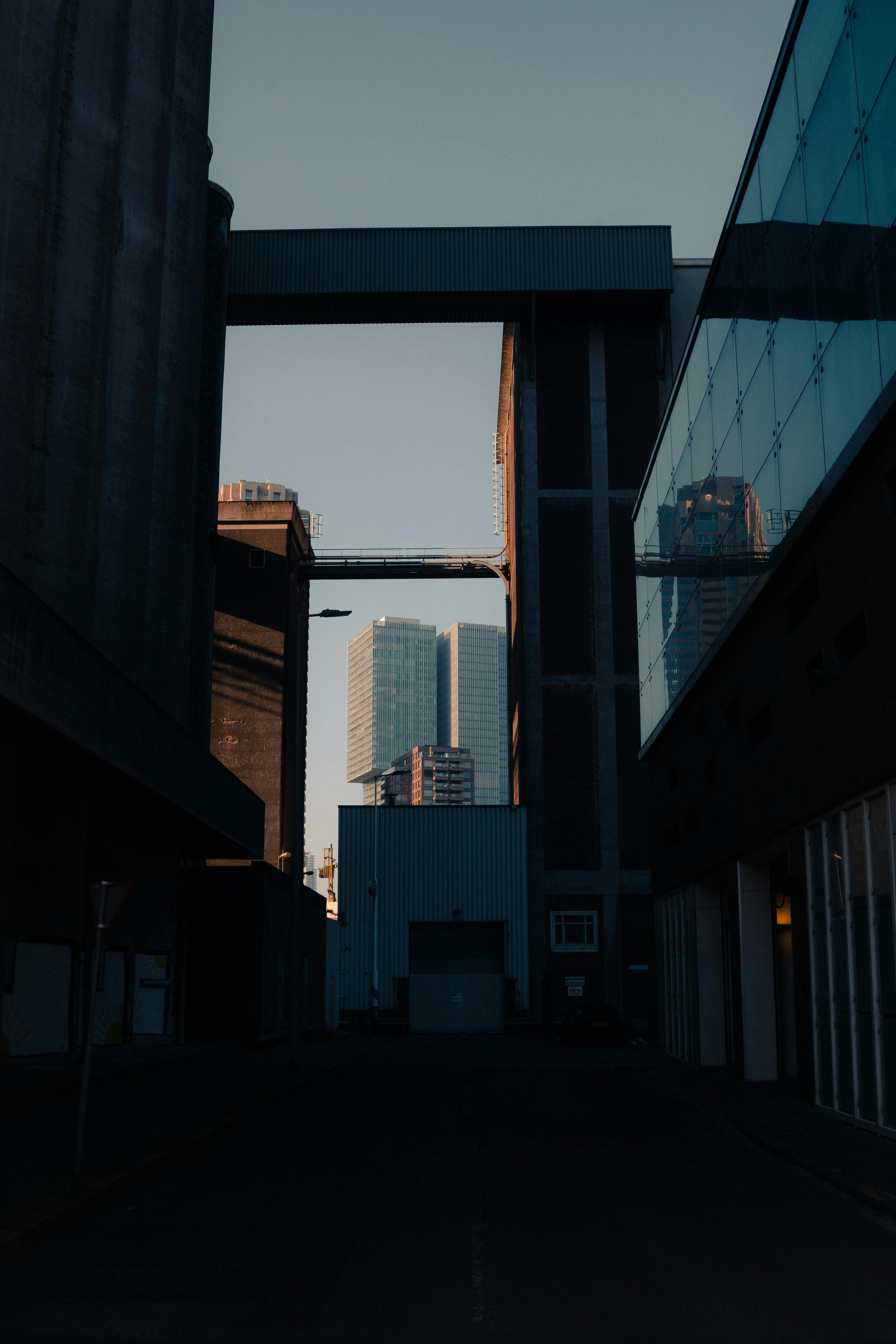 A narrow urban alleyway at dusk, framed by tall industrial buildings, with modern skyscrapers visible in the background.