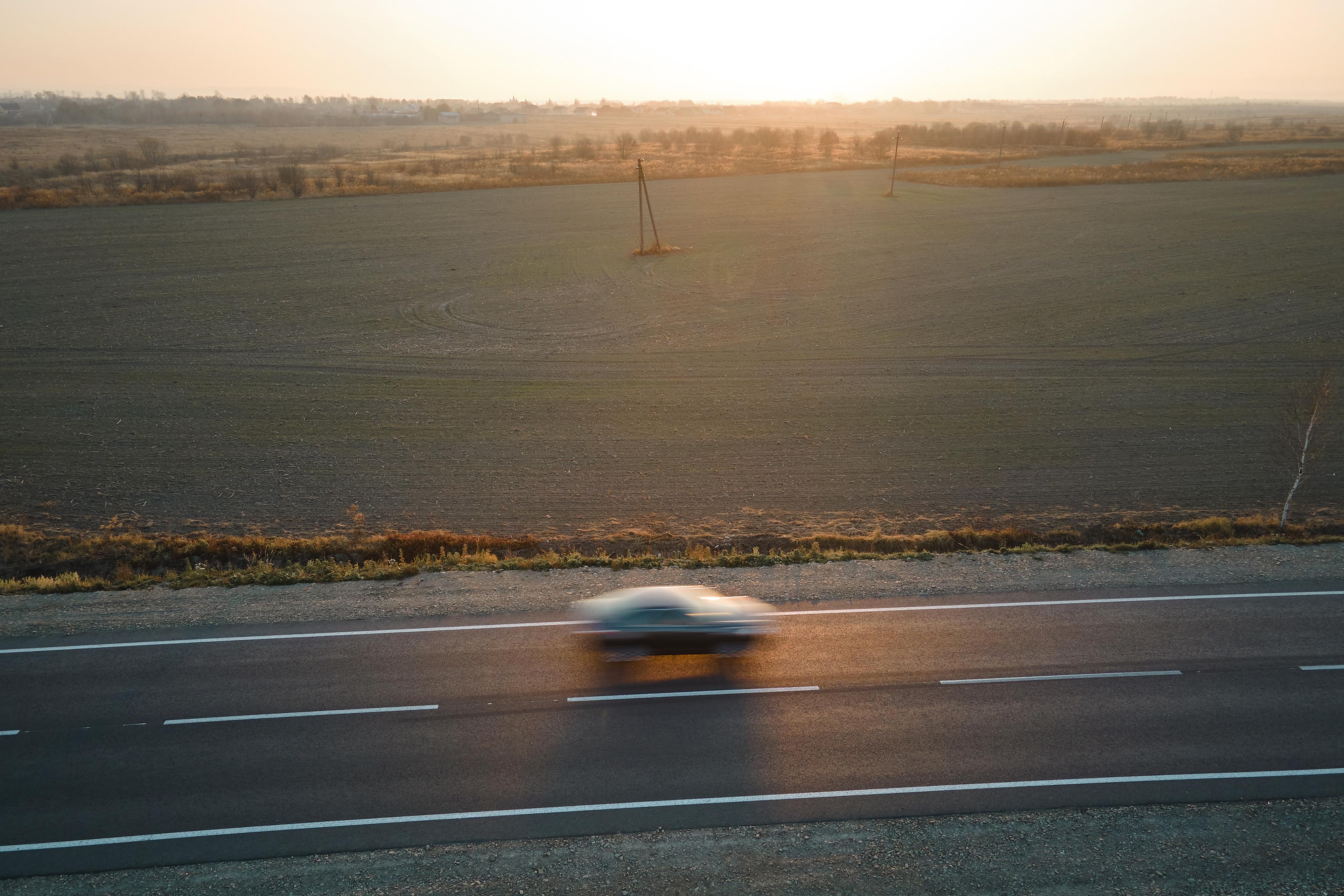 Aerial view of a car speeding on a rural road at sunset, with vast open fields and distant trees in the background.