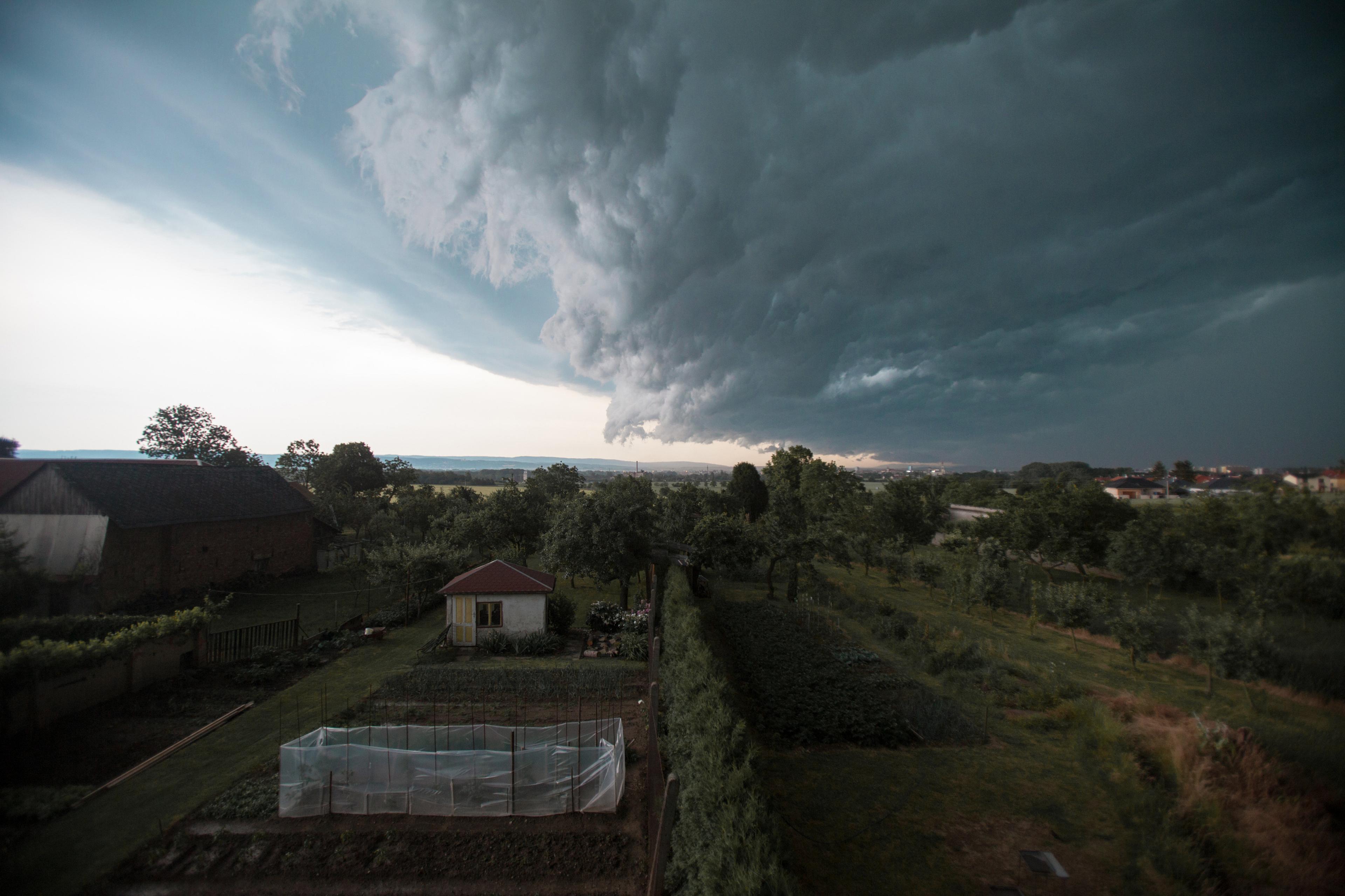 A storm approaches a rural landscape with dramatic, dark clouds covering the sky, casting shadows over fields and a small garden.