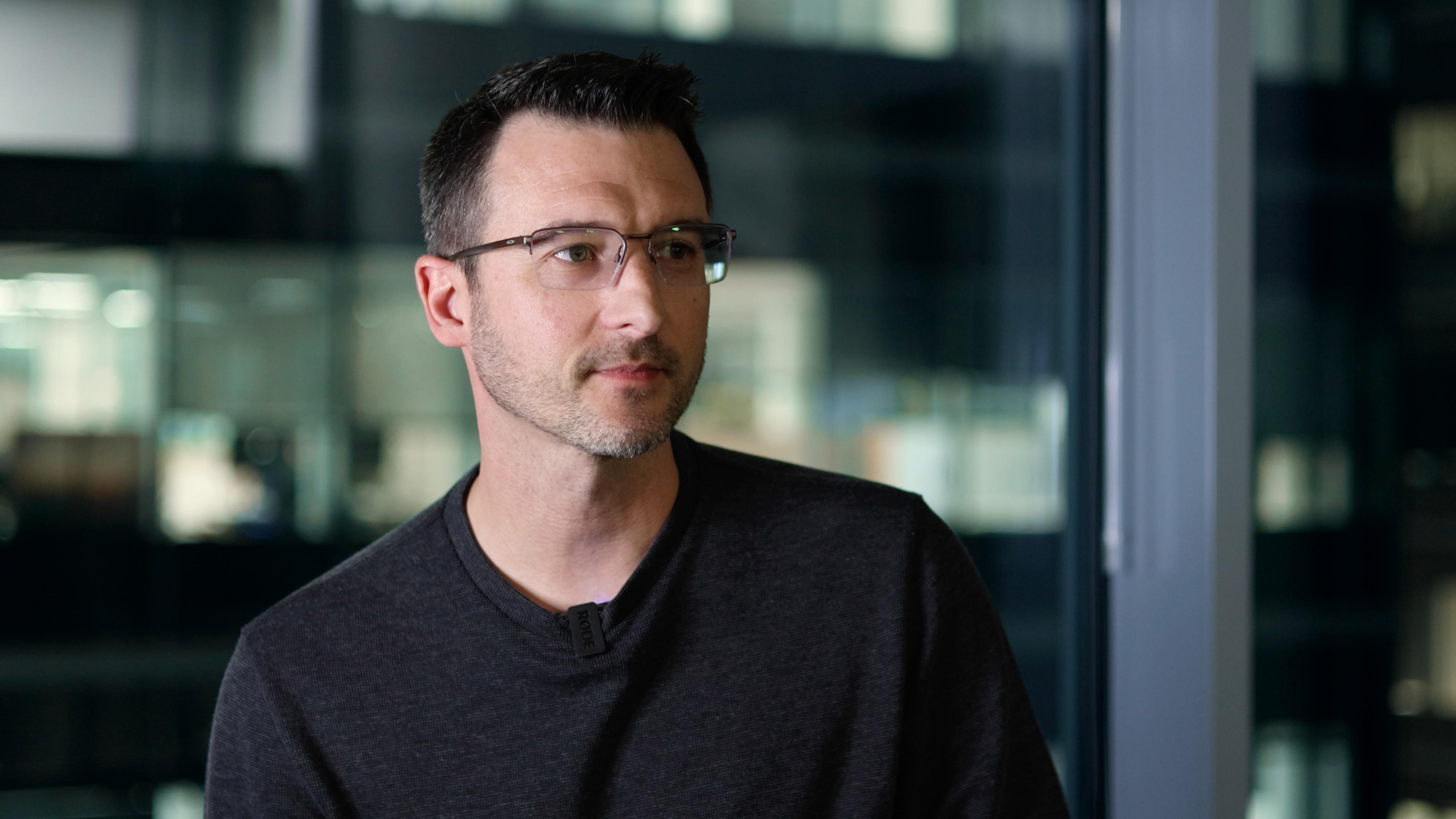 Scott Mackaro, a man with glasses and short hair wearing a dark sweater sits in front of a large window with blurred office background.