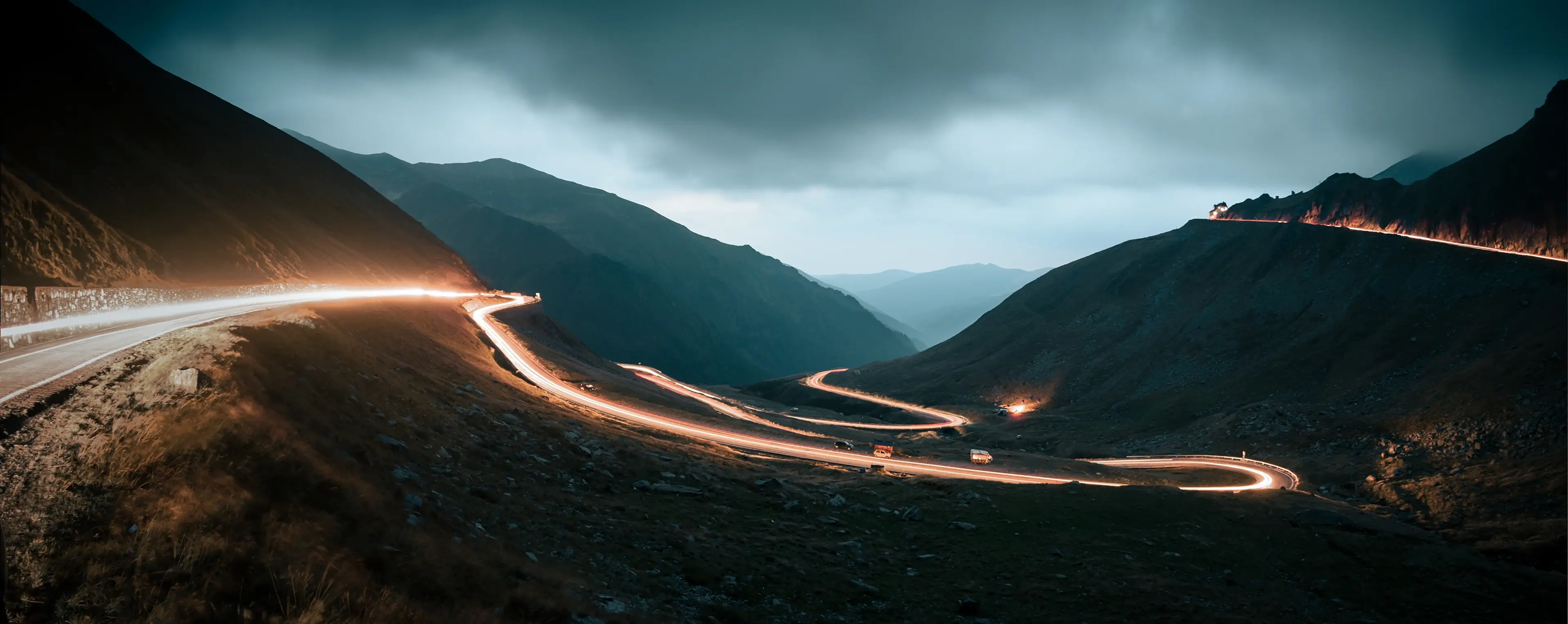 Winding mountain road at night with bright car light trails and a dark, cloudy sky in the background.