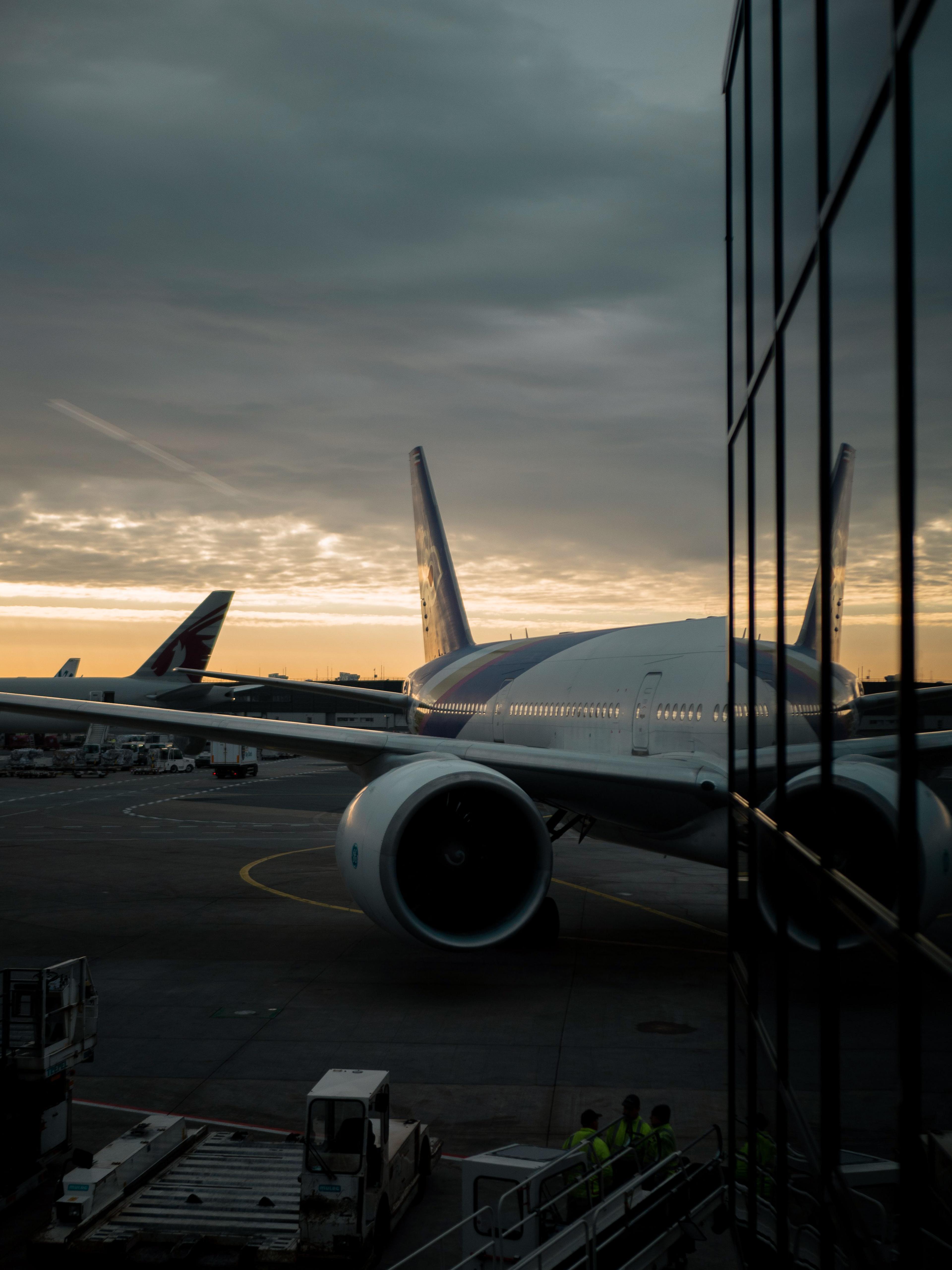 A large airplane is parked at an airport gate during sunset, with a cloudy sky reflected in nearby glass windows. Workers are near the aircraft.