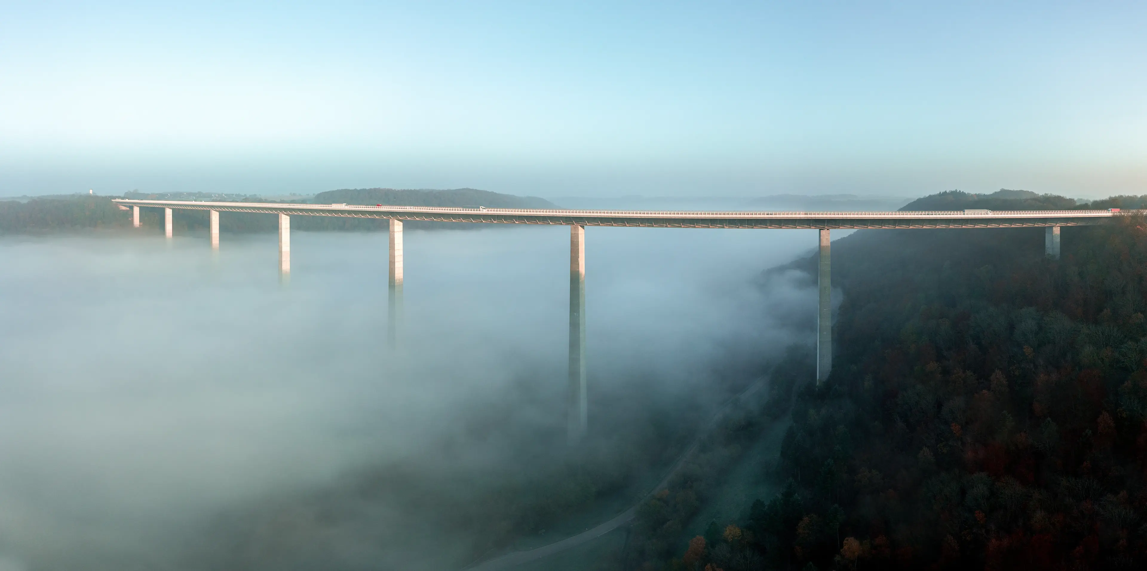 High bridge stretches over a fog-filled valley, supported by tall pillars, with dense forest below and clear sky above.
